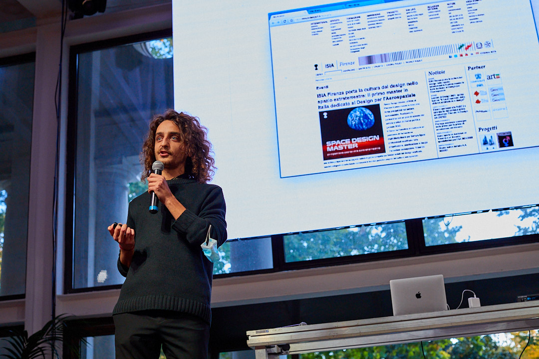 Persona che parla davanti a un pubblico con microfono, slide su grande schermo, presentazione su design spaziale.