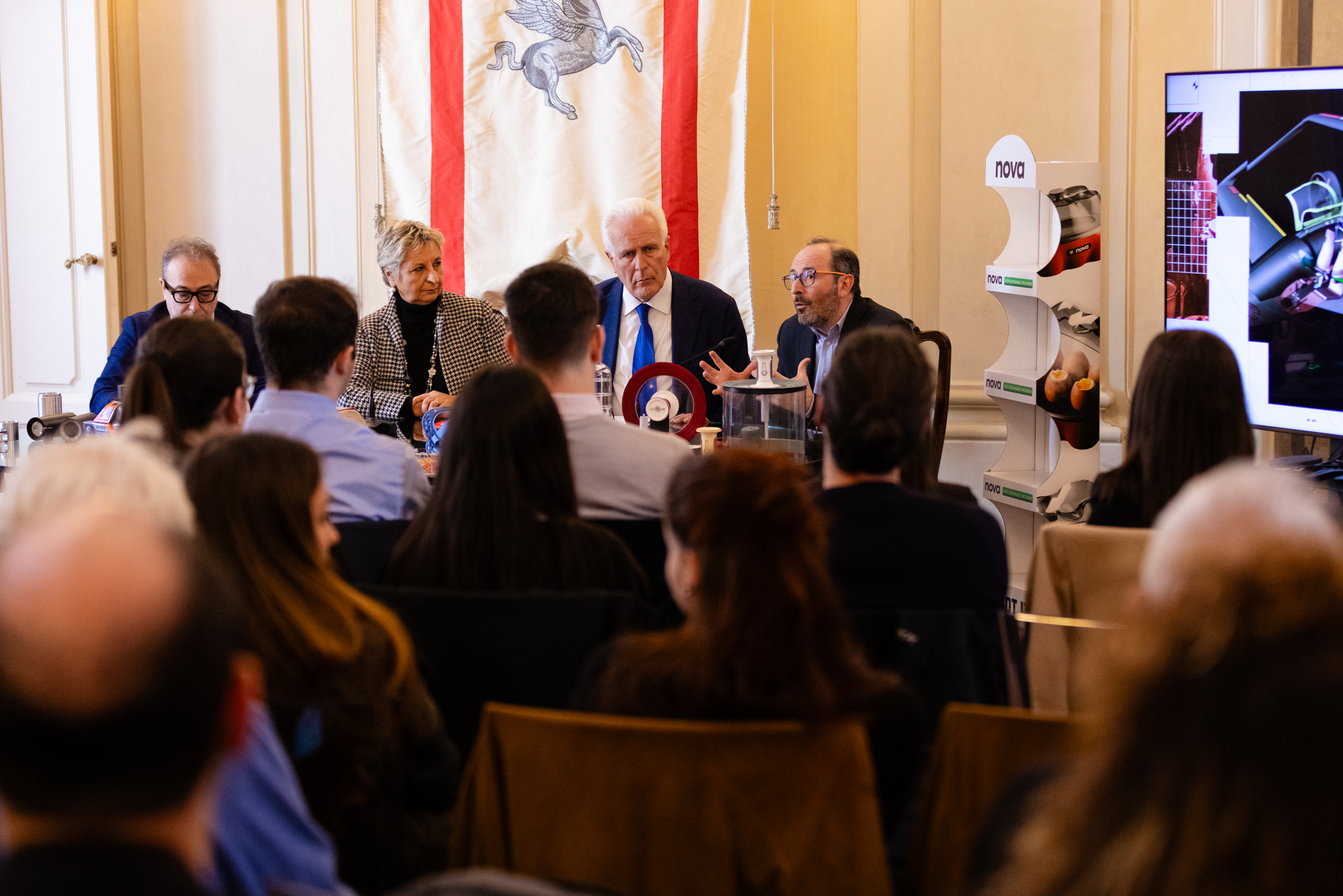 Conferenza stampa con quattro relatori seduti a un tavolo, pubblico e schermo laterale in una sala formale.