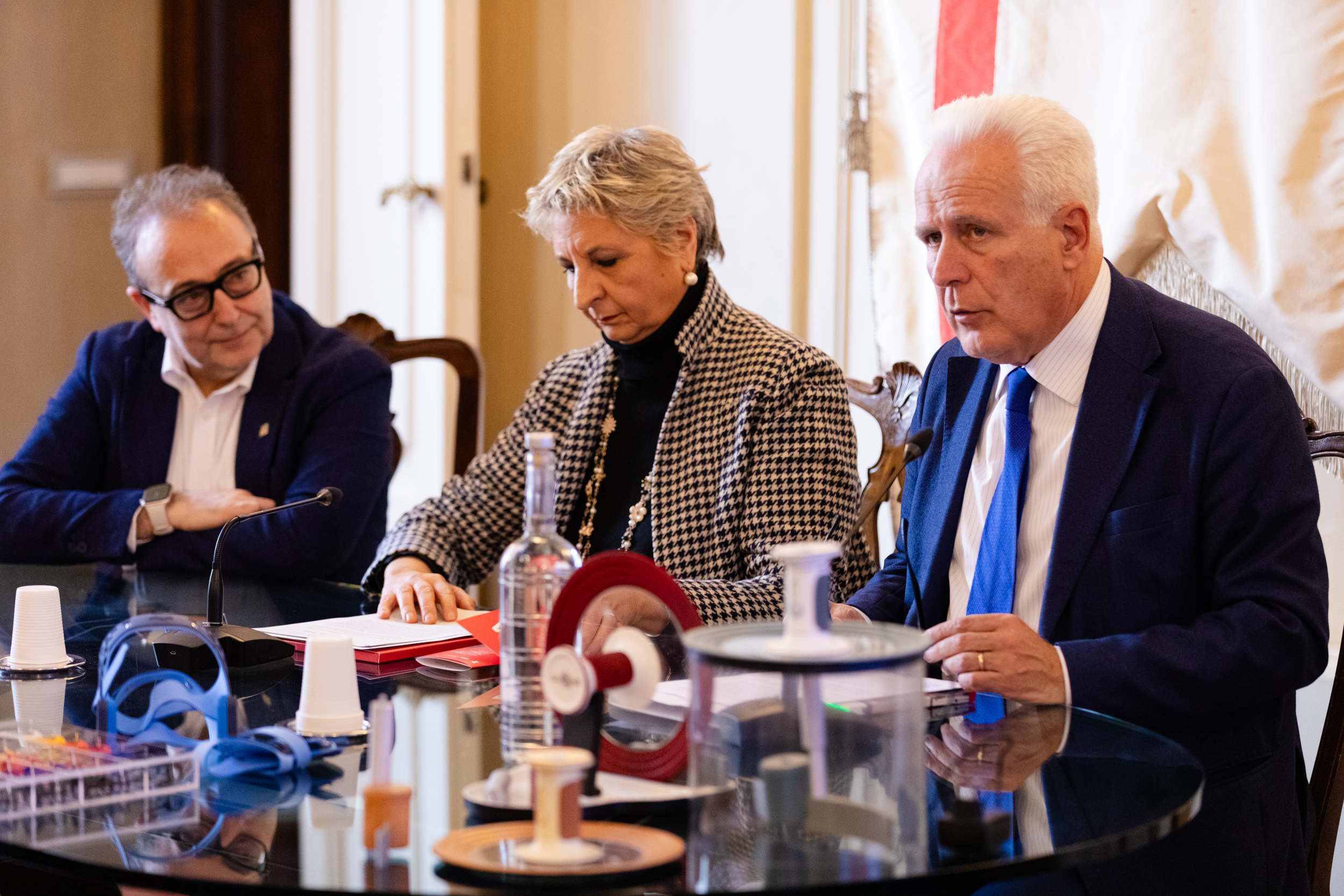 Tre persone siedono a un tavolo da riunione con documenti e attrezzature scientifiche in primo piano.