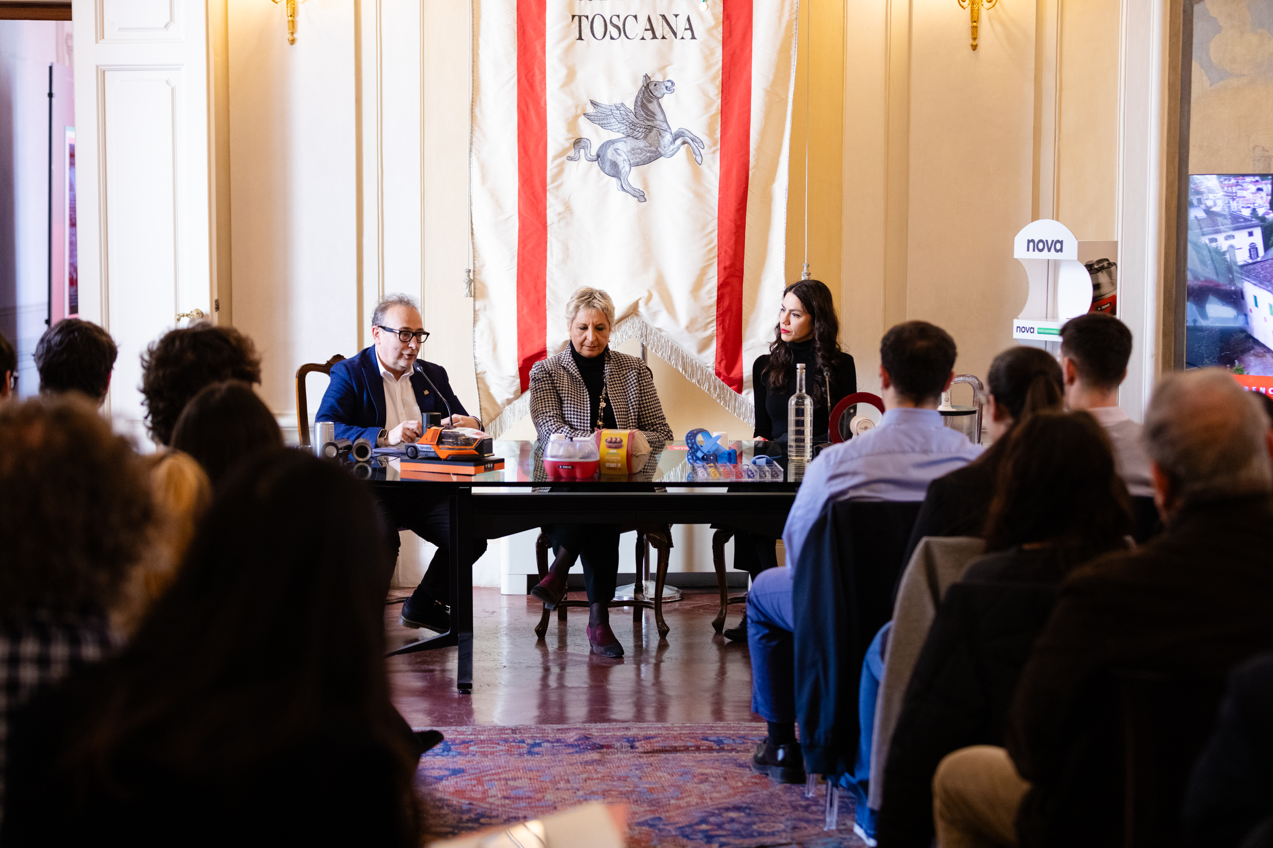 Conferenza in Toscana con tre relatori seduti a un tavolo e pubblico attento in una sala elegante.