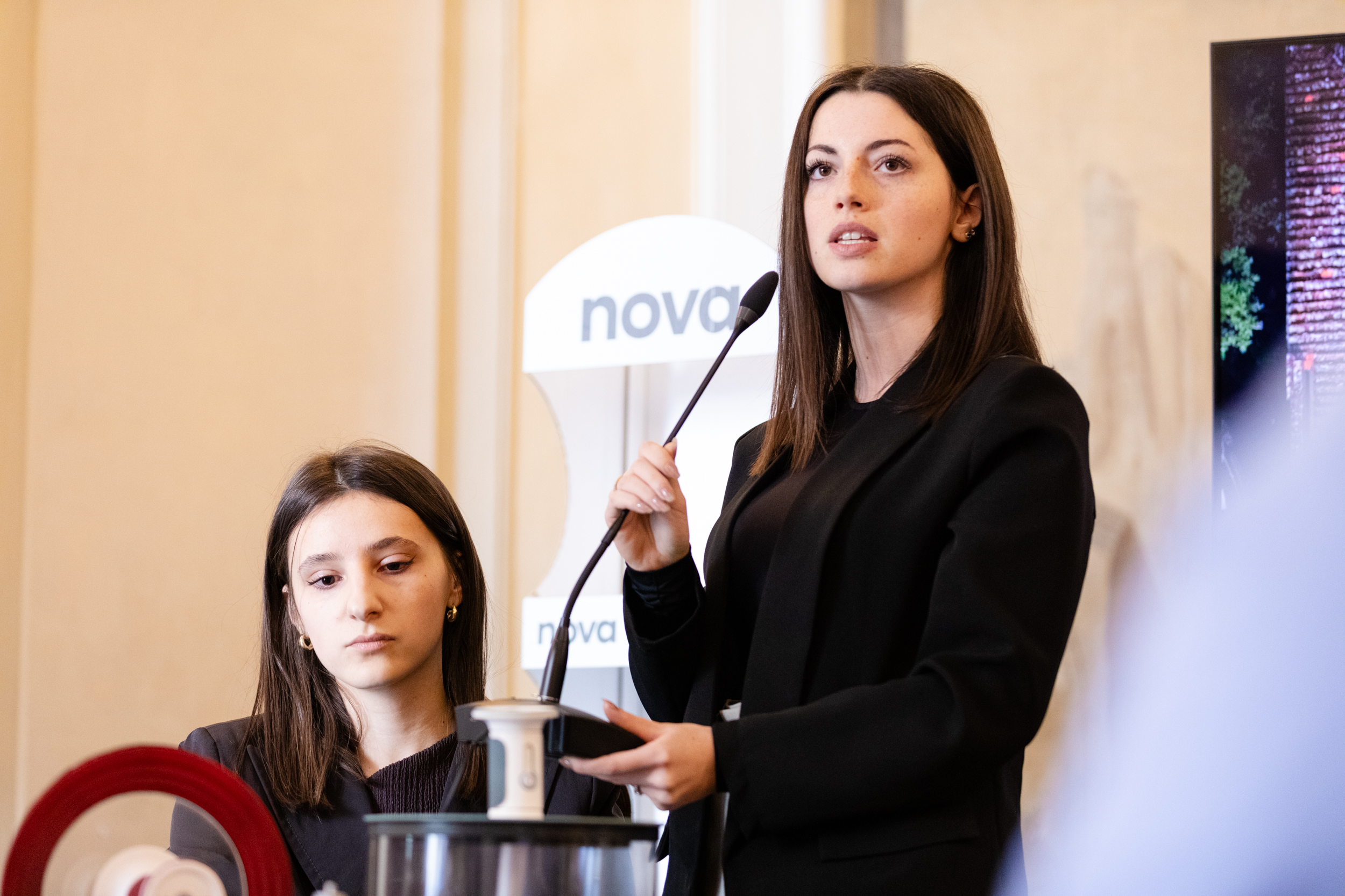 Due donne in abiti eleganti partecipano a una conferenza, una parla al microfono, l'altra è concentrata.