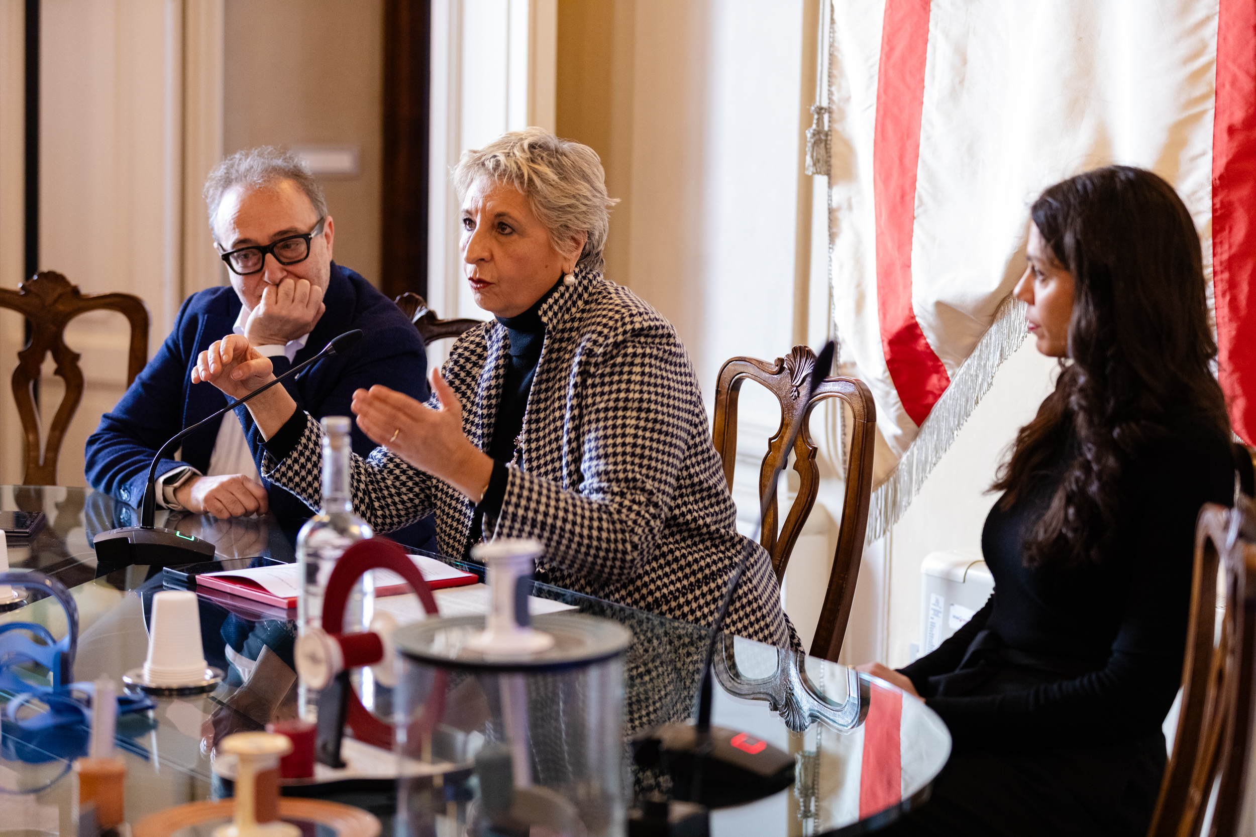 Tre persone discutono sedute a un tavolo di vetro durante una conferenza in una sala elegante con bandiera sullo sfondo.