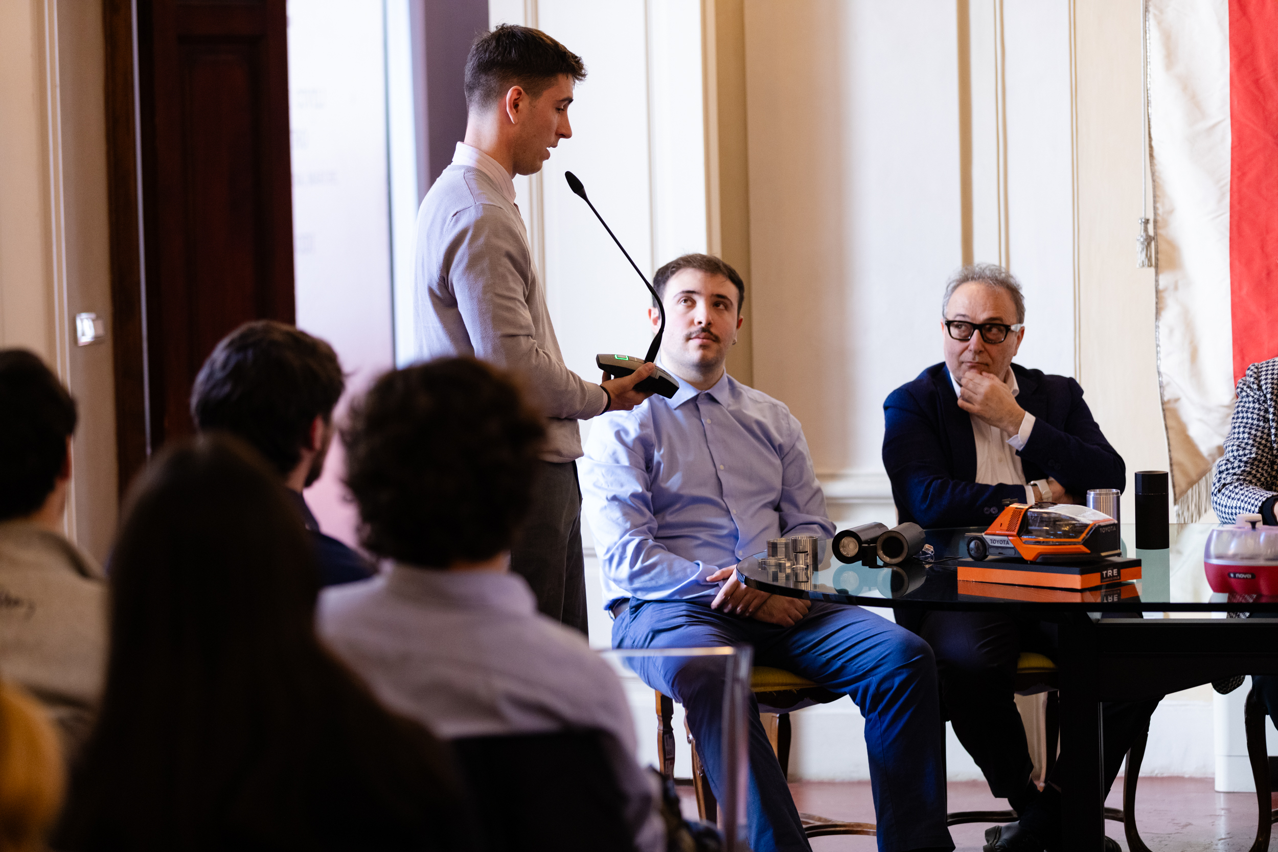 Uomo parla a una conferenza, pubblico ascolta. Tavolo con oggetti tecnologici sullo sfondo. Ambiente formale e serio.