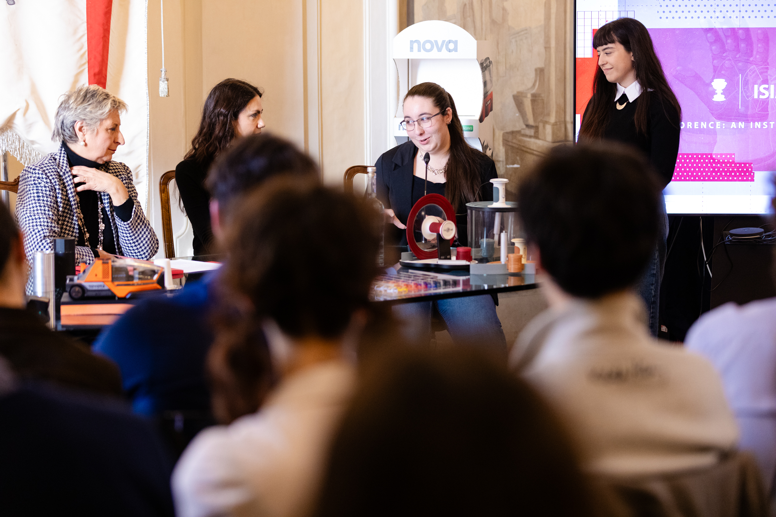 Un gruppo di persone discute animatamente durante una conferenza, con schermo e attrezzature tecniche sul tavolo.