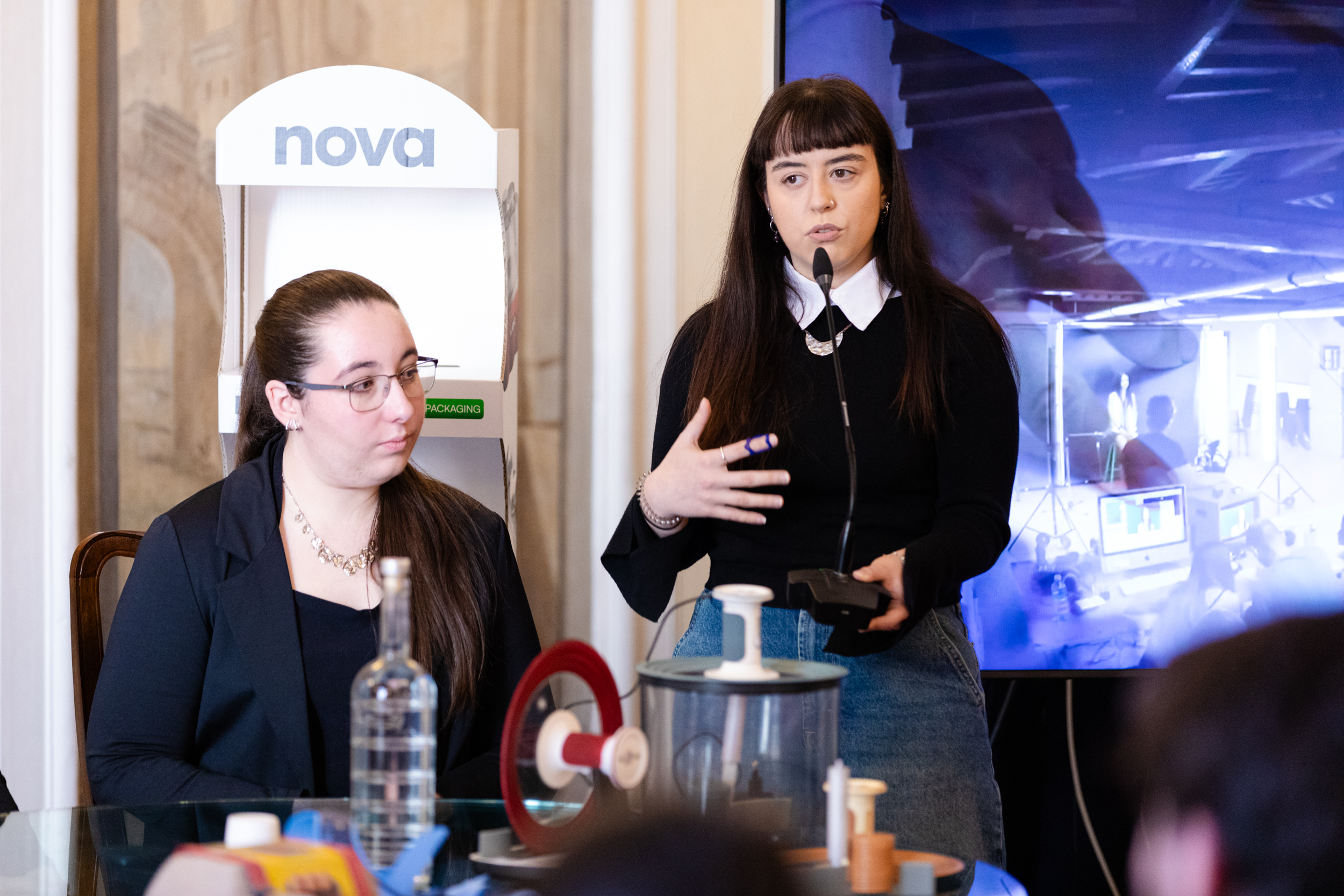 Due donne partecipano a una presentazione tecnologica in un ambiente formale con attrezzature sul tavolo.