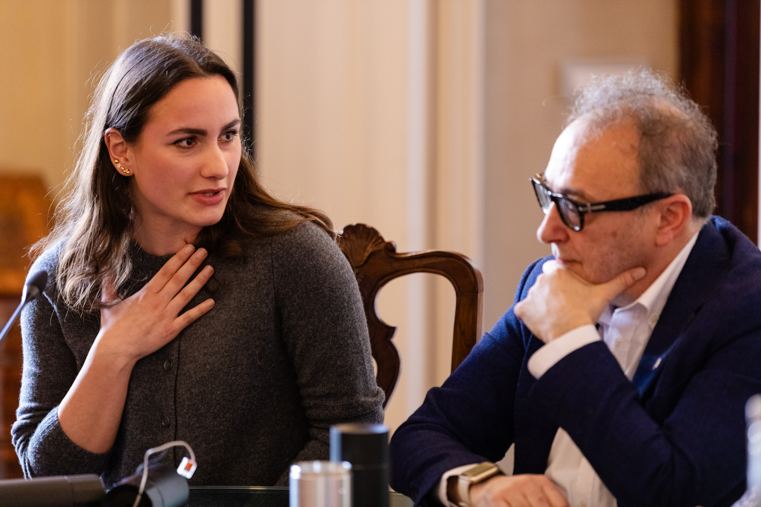 Due persone conversano in una sala conferenze, sedute a un tavolo, in un contesto formale ed elegante.