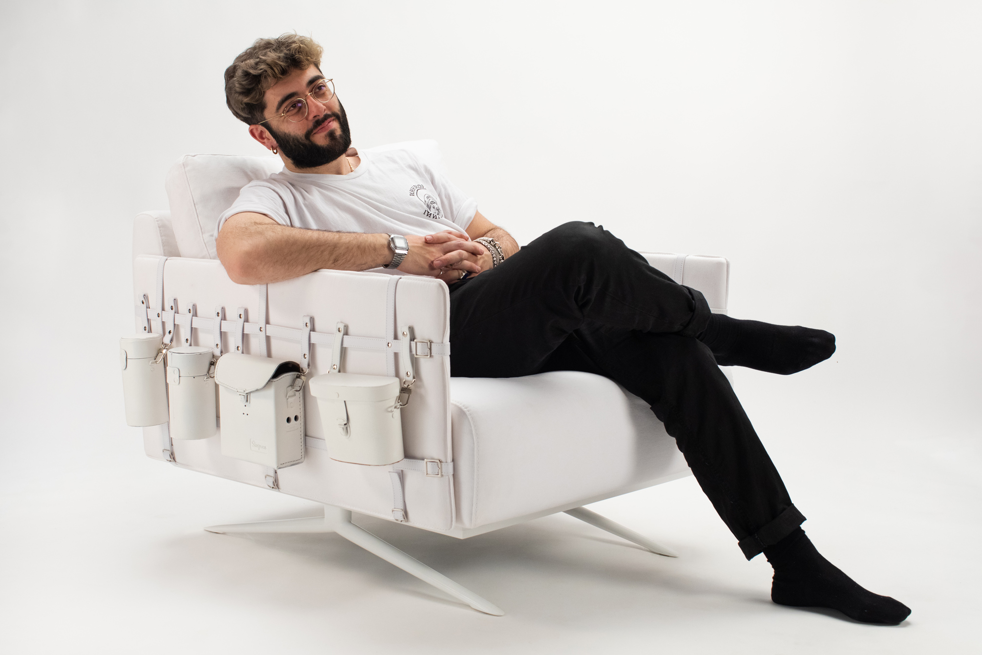 Uomo seduto rilassato su poltrona bianca con tasche, indossa occhiali, maglietta bianca e pantaloni neri.