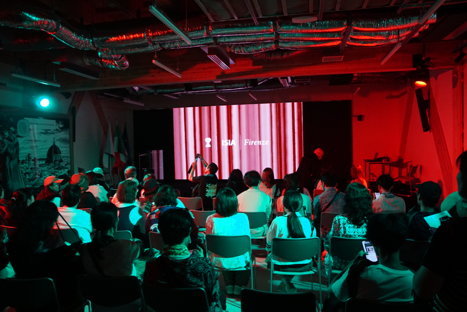 Persone sedute assistono a una presentazione in un auditorium dell'ISIA Firenze con luci colorate.