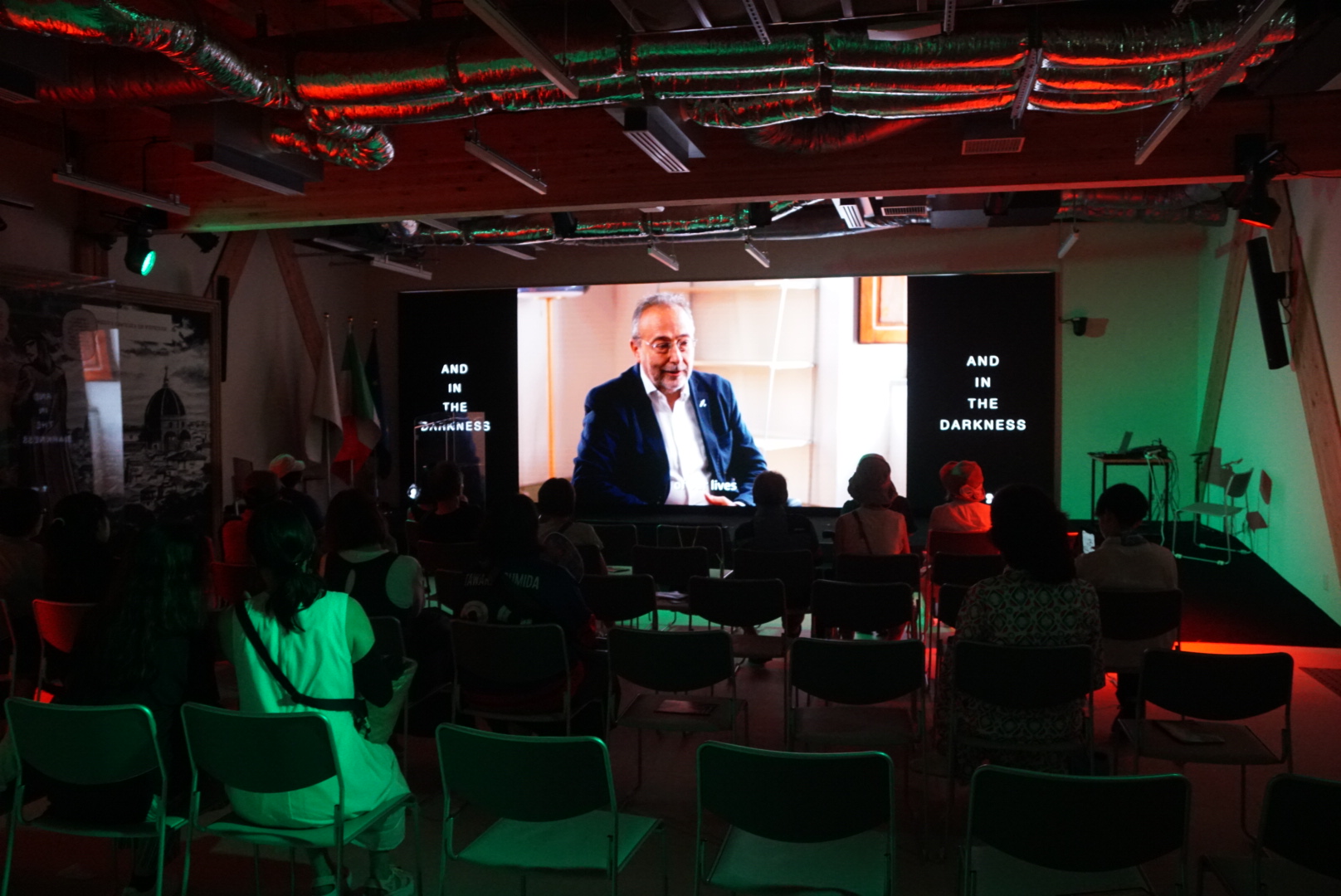 Persone sedute guardano un video proiettato in sala con illuminazione soffusa, bandiere italiane visibili.