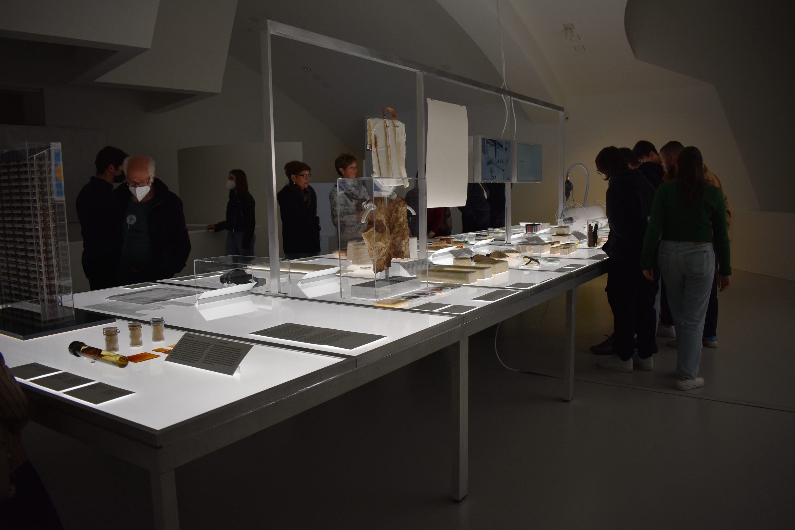 Visitatori osservano un'esposizione di arte e cultura moderna in un museo, analizzando dettagli e oggetti esposti su un tavolo.