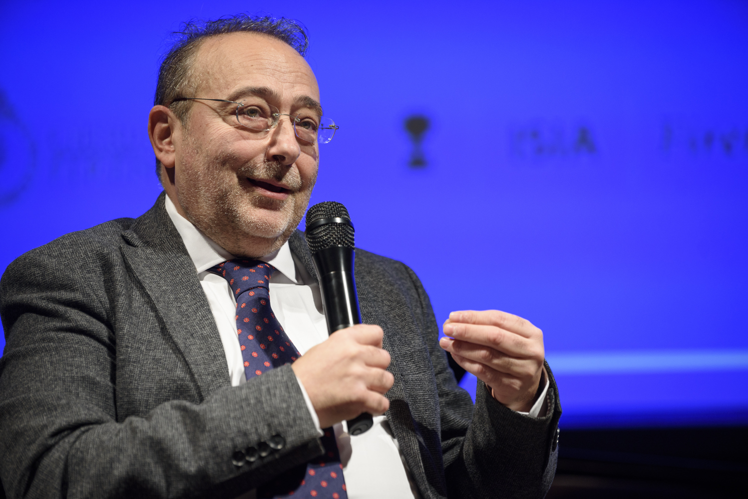 Persona che parla al microfono su sfondo blu durante un evento.