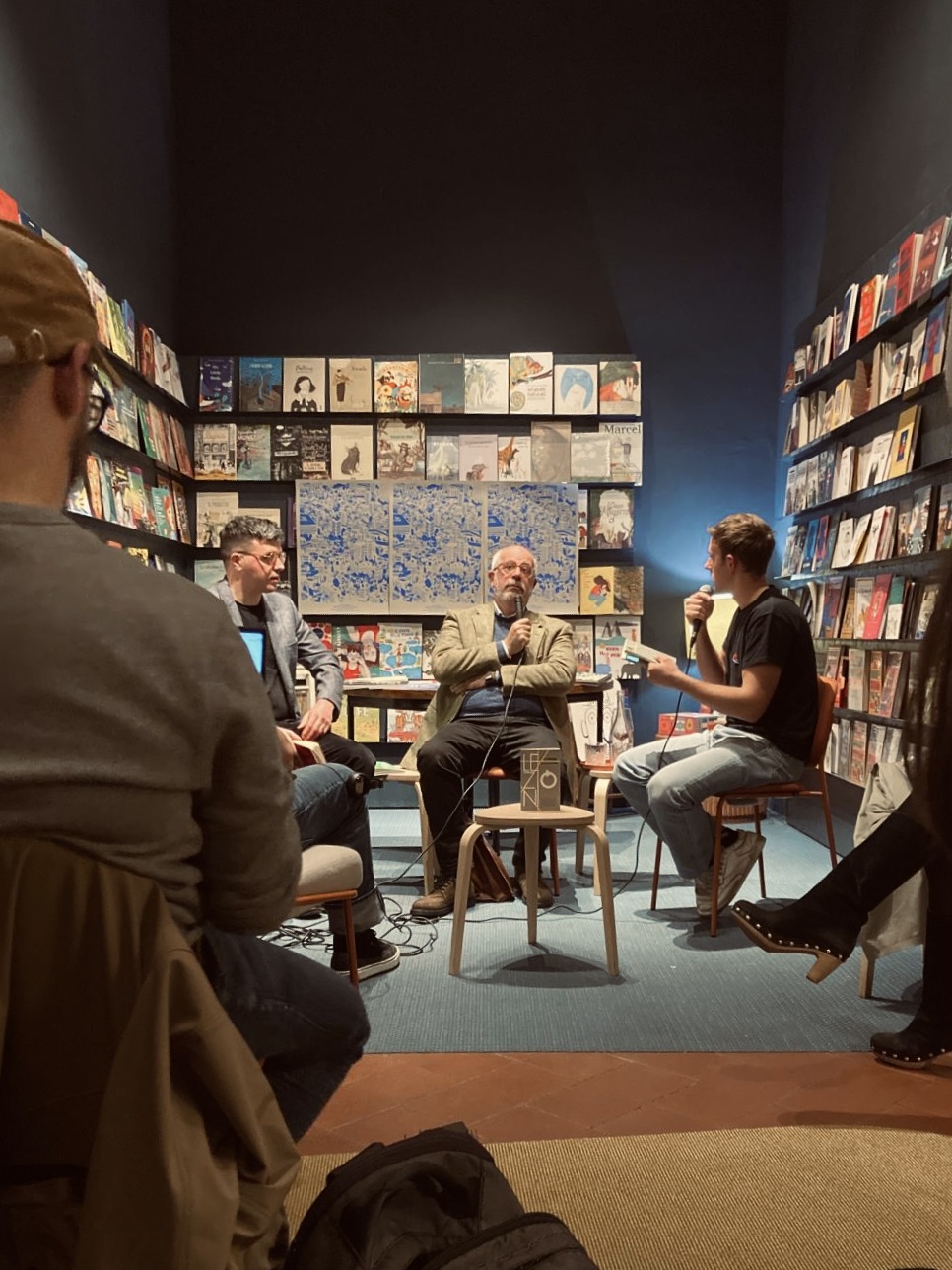 Incontro letterario in libreria con conversazione tra tre uomini seduti, circondati da scaffali pieni di libri.