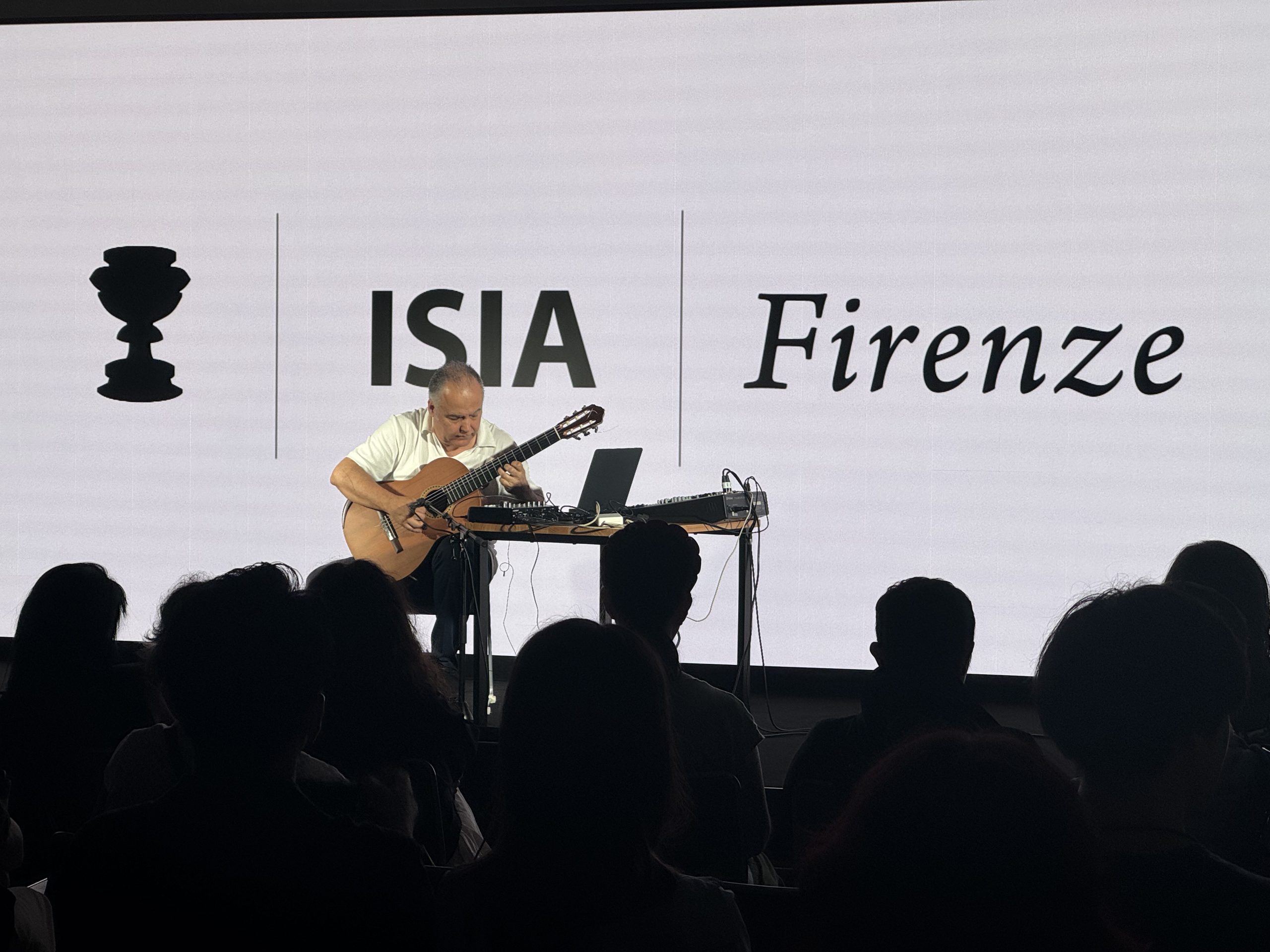 Musicista suona la chitarra su un palco all'evento ISIA Firenze, pubblico seduto in silenzio.