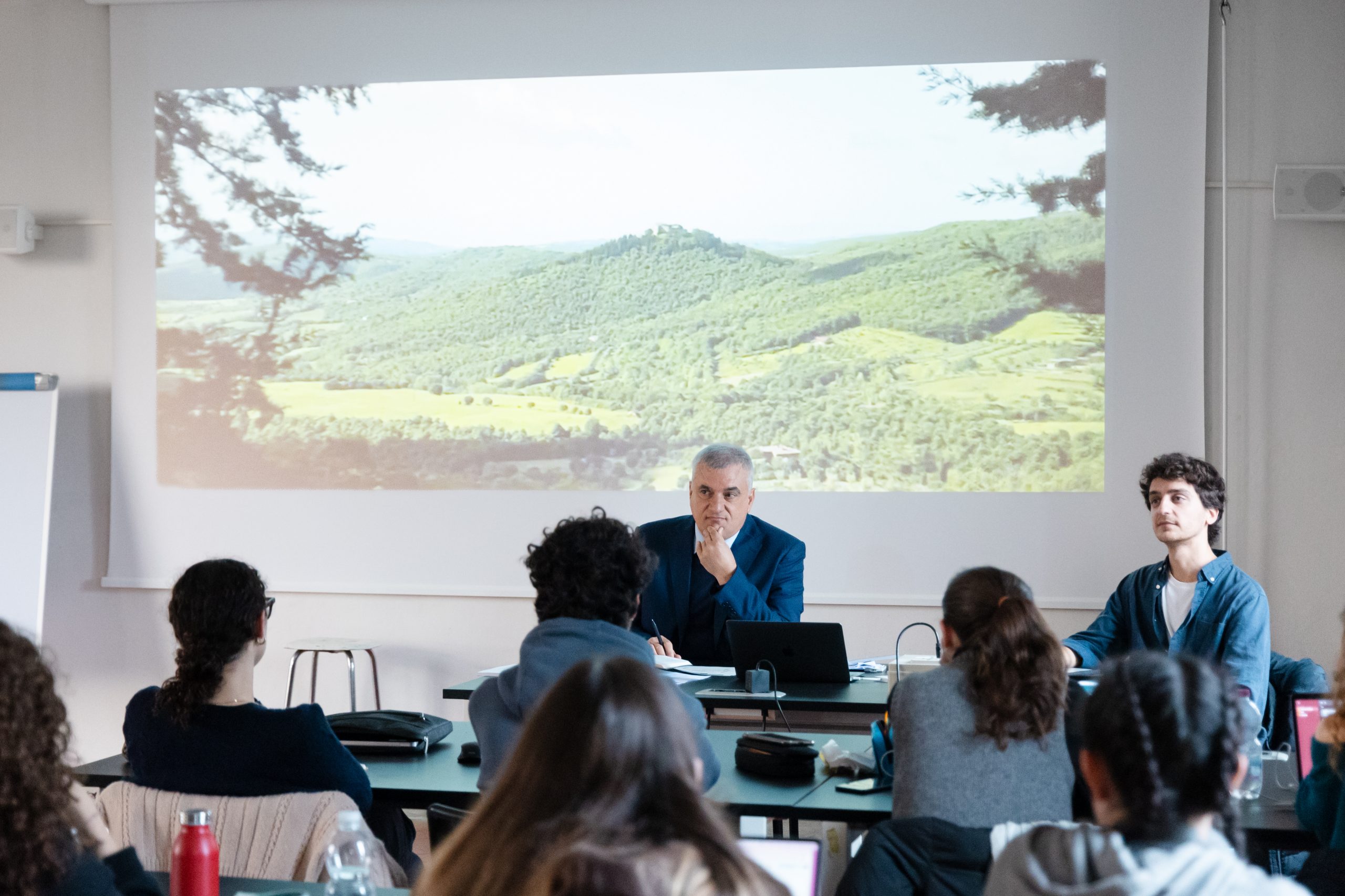 Una lezione in aula con un relatore e una proiezione di paesaggio. Studenti ascoltano attentamente.