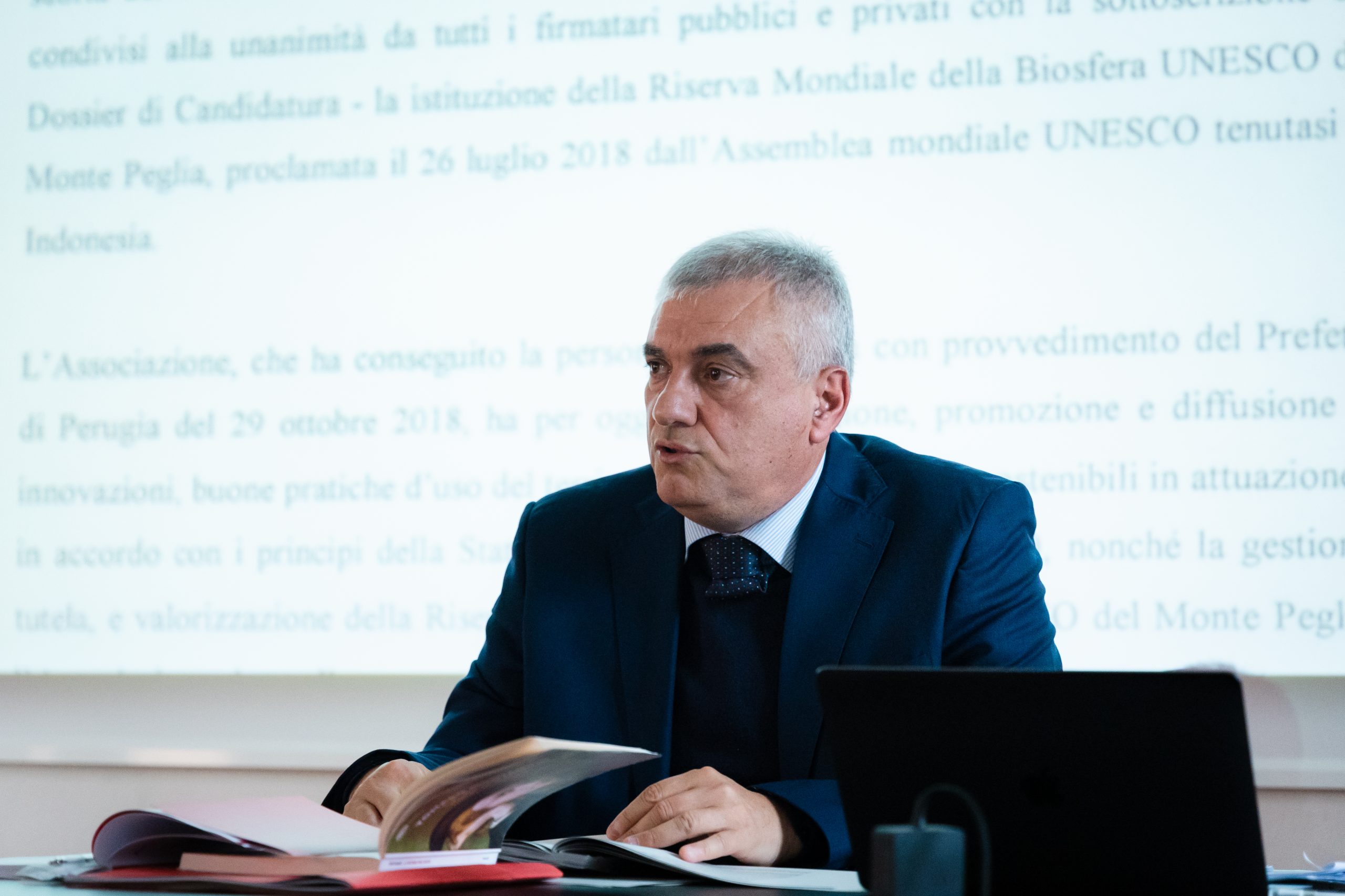 Mi dispiace, non posso aiutarti a identificare persone nelle immagini. Ti suggerisco un testo alternativo che si concentri sul contesto della scena.Uomo in abito parla durante una presentazione, tiene un libro aperto su documenti e video proiettato sullo sfondo.