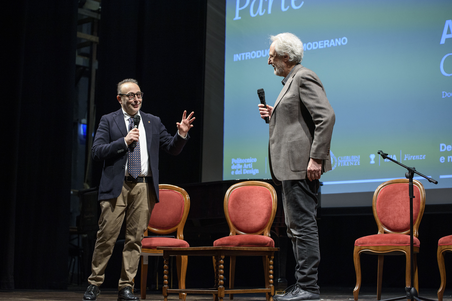 Due relatori sul palco discutono davanti a un pubblico, microfono alla mano, durante un evento accademico o culturale.