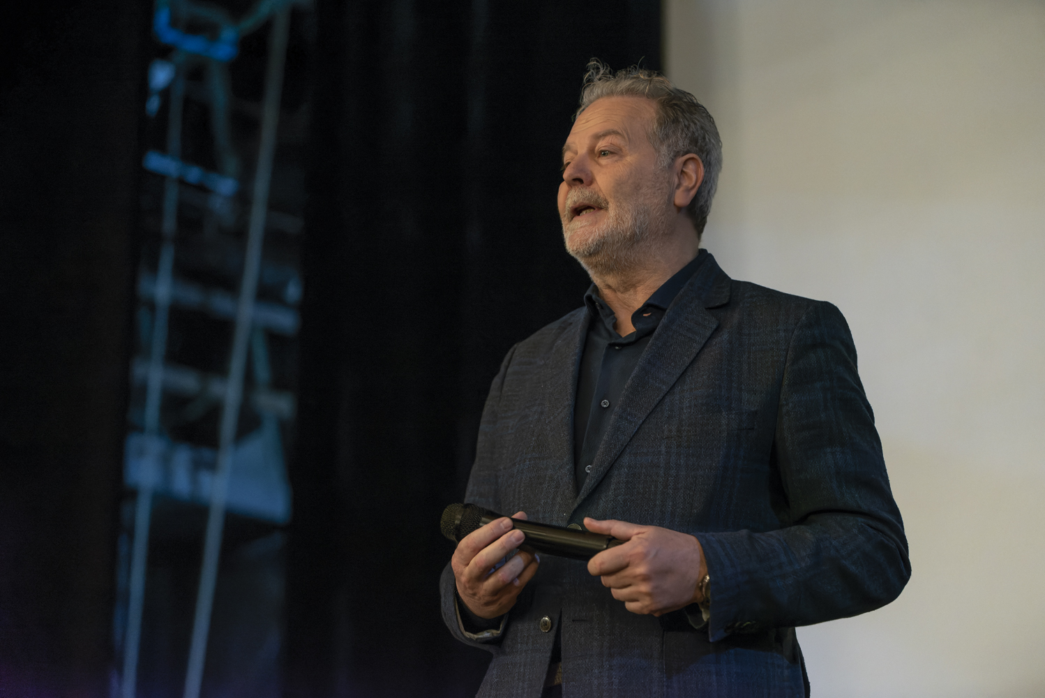 Uomo con microfono in mano, in giacca blu scuro, parla su un palco con sfondo neutro.