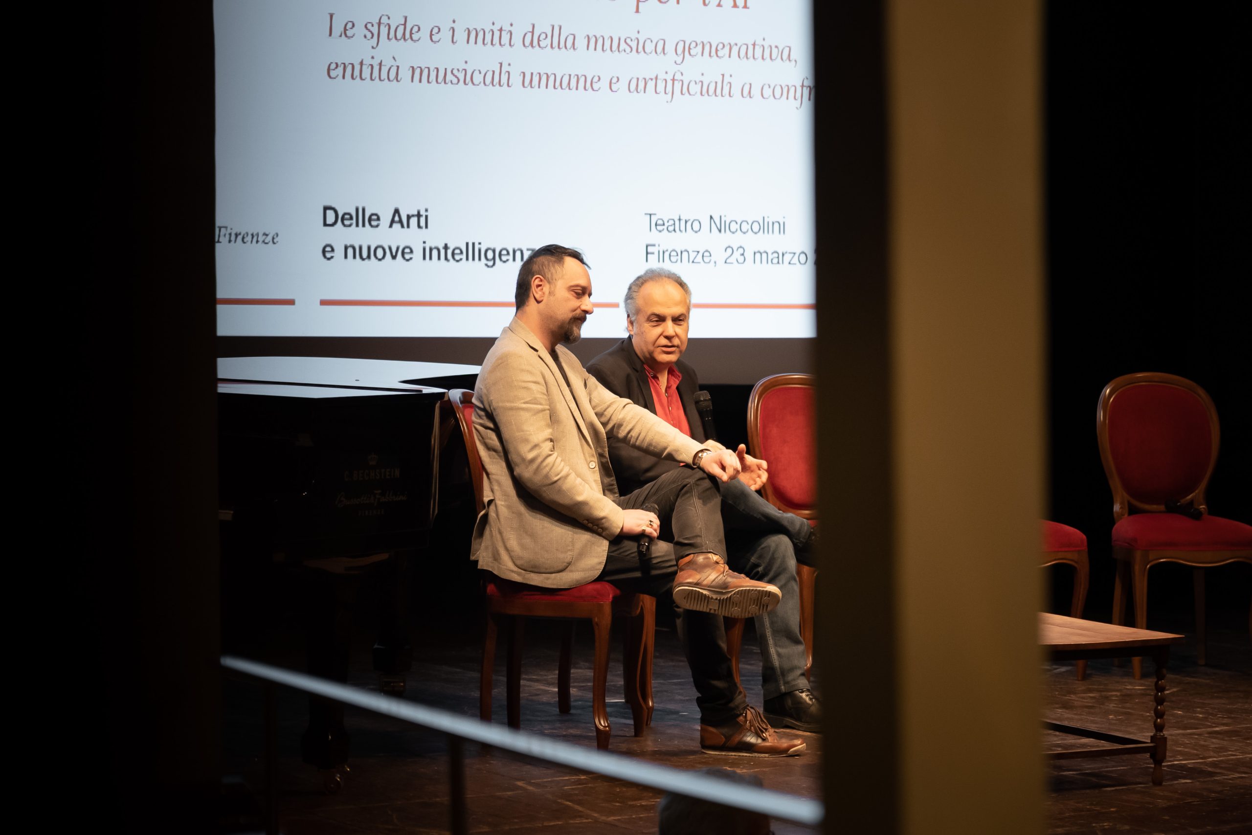 Due uomini seduti su un palco discutono di musica generativa al Teatro Niccolini di Firenze, evento del 23 marzo.