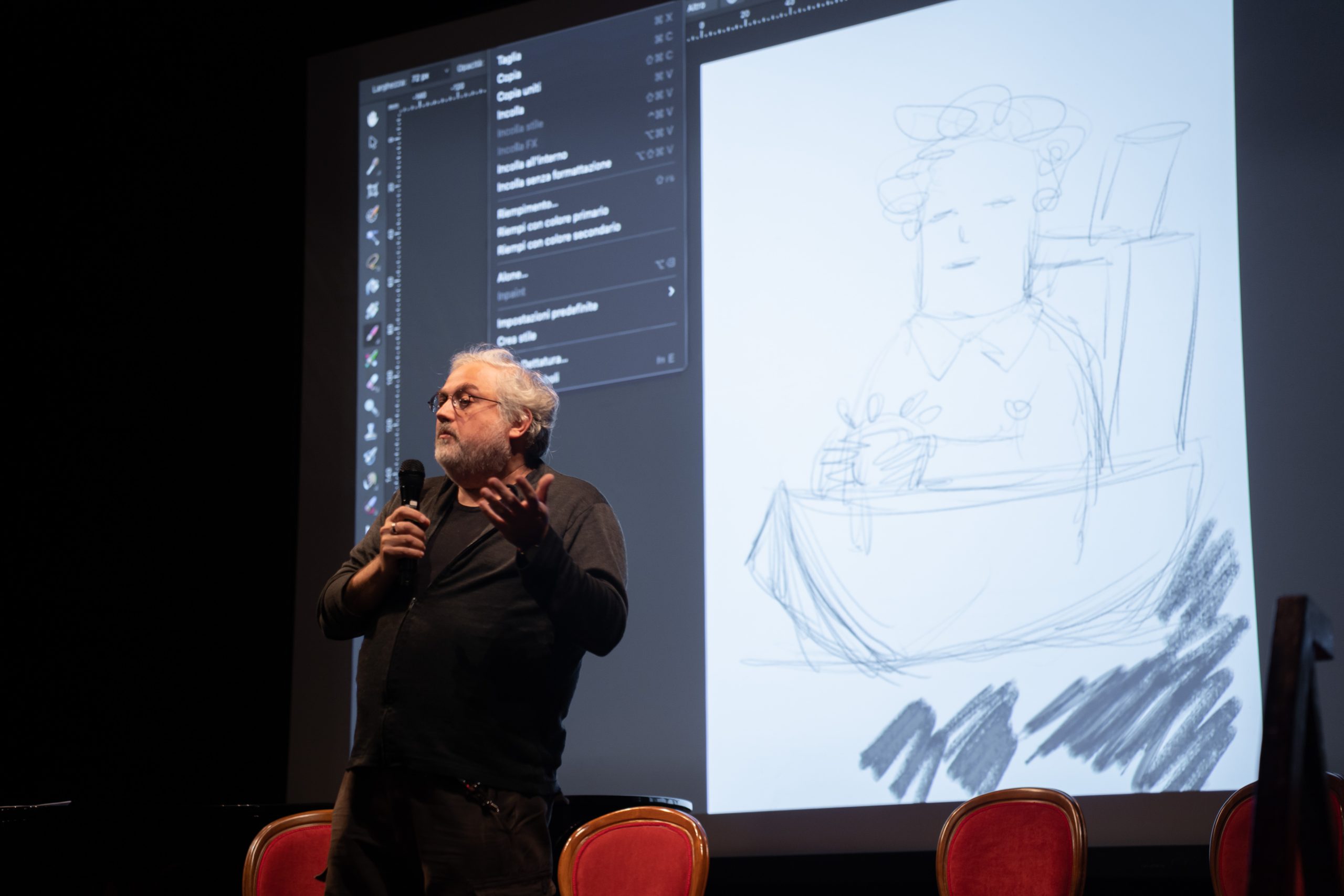 Uomo che parla al microfono con disegno proiettato su schermo dietro di lui, durante una presentazione.