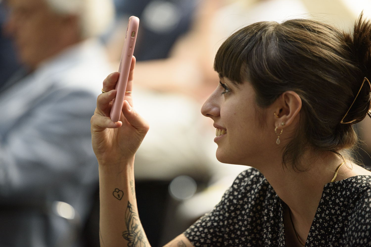Donna sorridente con smartphone rosa, scatta una foto in un ambiente interno durante un evento pubblico.