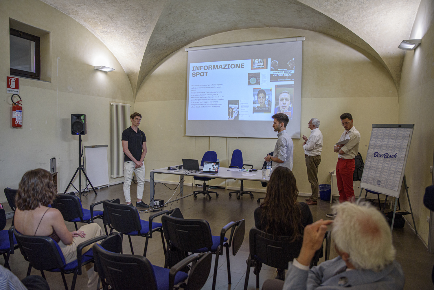 Presentazione in aula con pubblico seduto e proiezione su schermo di diapositive informative.