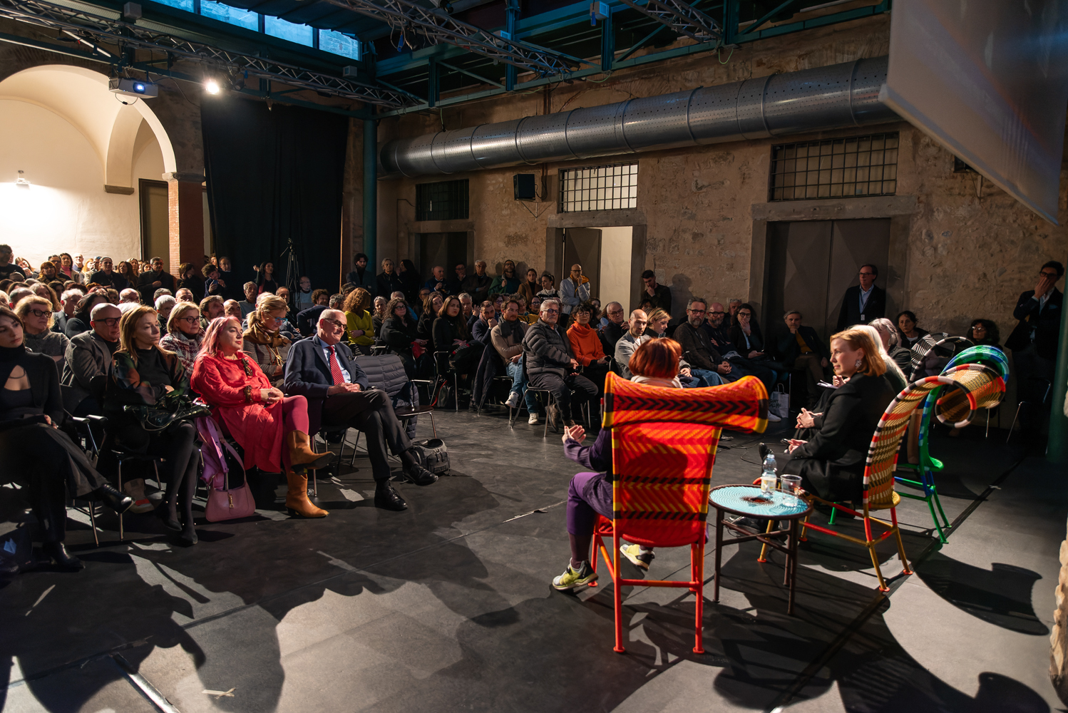 Discussione e pubblico attento in un evento culturale in uno spazio industriale con sedie colorate.