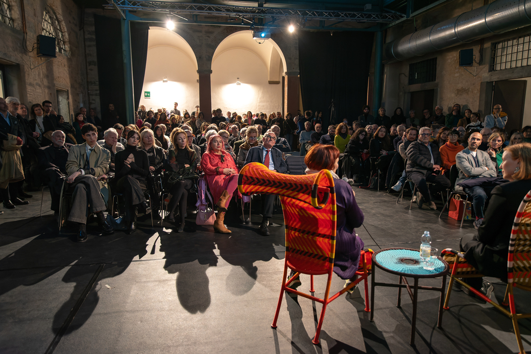 Pubblico attento a un evento culturale, seduto in una sala con relatori su sedie colorate in primo piano.