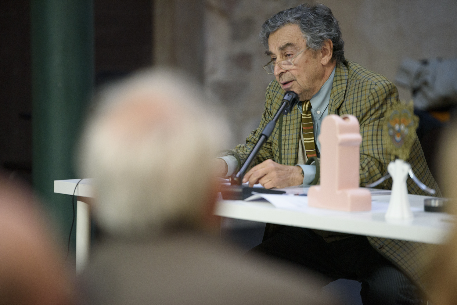 Uomo anziano in giacca a quadri parla al microfono durante una conferenza, seduto a un tavolo bianco con oggetti.