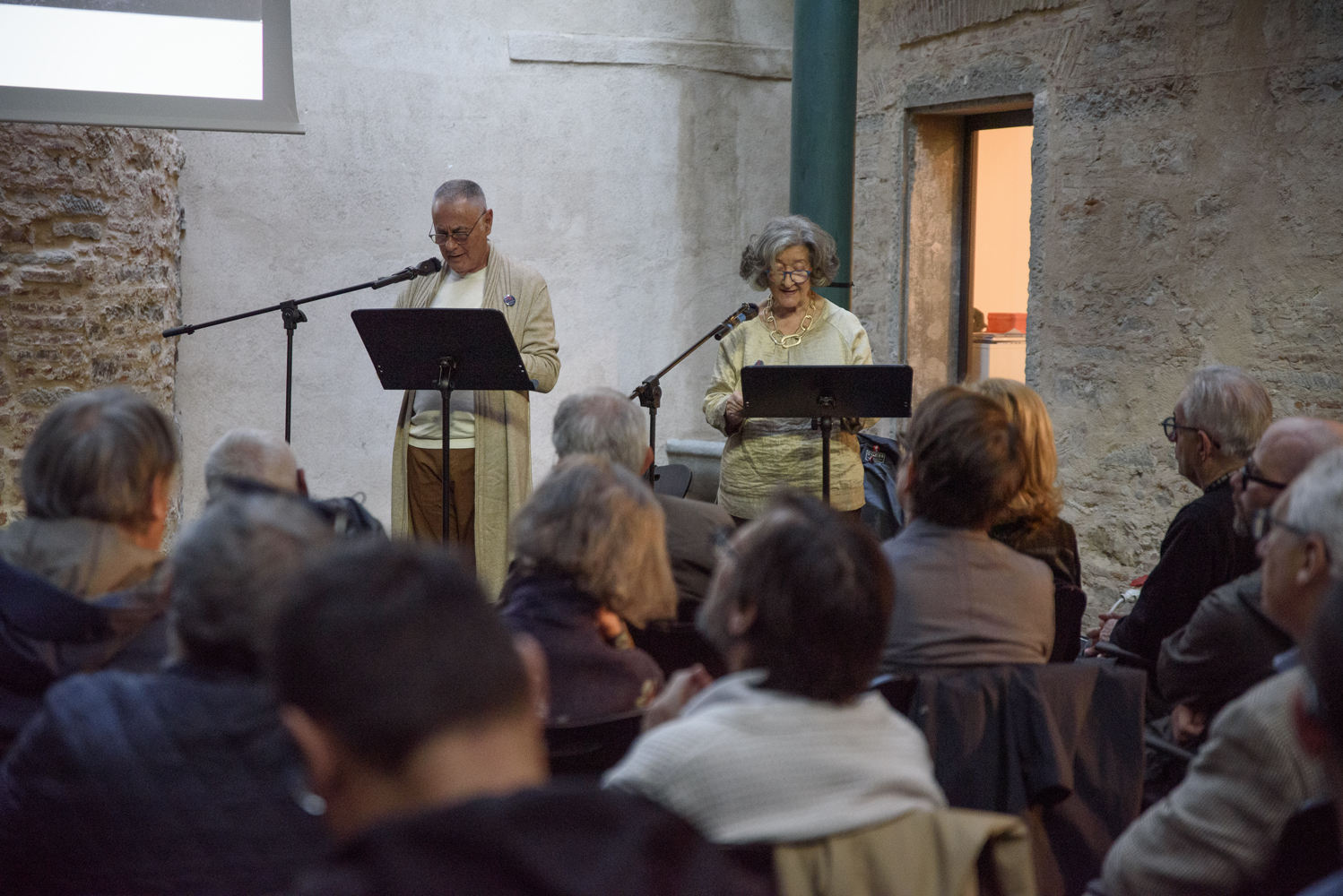 Due persone leggono al pubblico in un evento culturale in un ambiente storico con pareti di pietra.