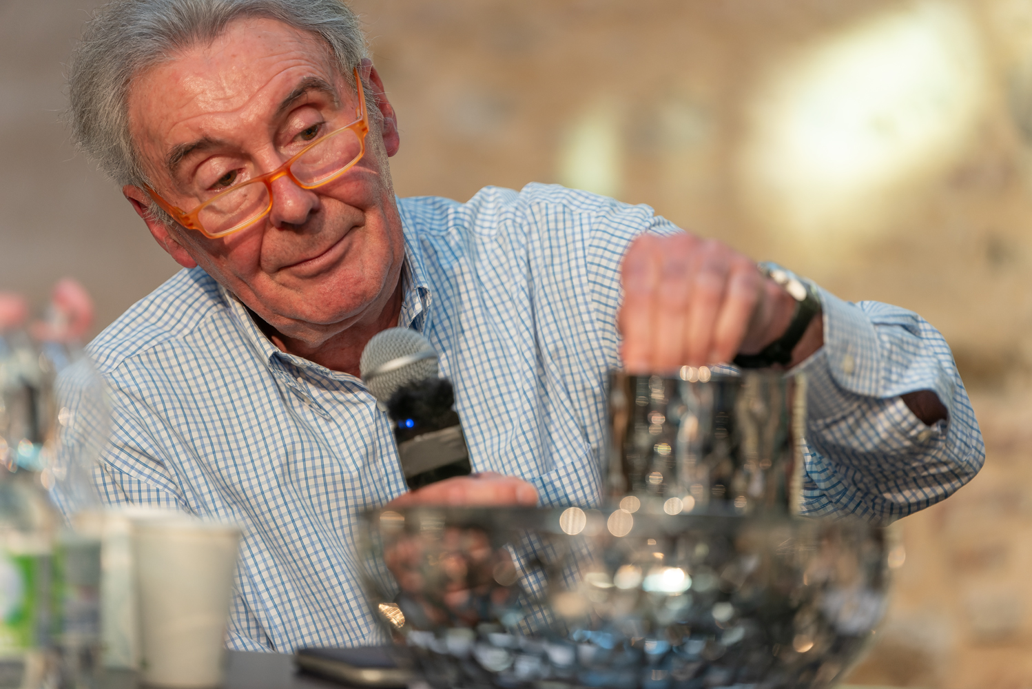 Uomo con occhiali arancioni, microfono in mano, in piedi davanti a un contenitore di cristallo su un tavolo.