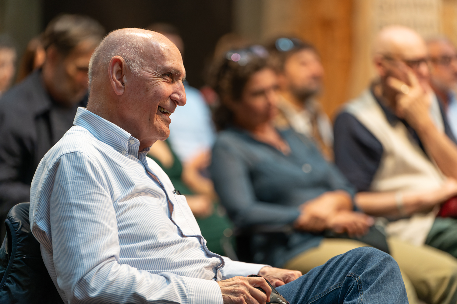 Uomo sorridente assiste a una conferenza, circondato da altre persone sedute, in un ambiente accogliente e informale.