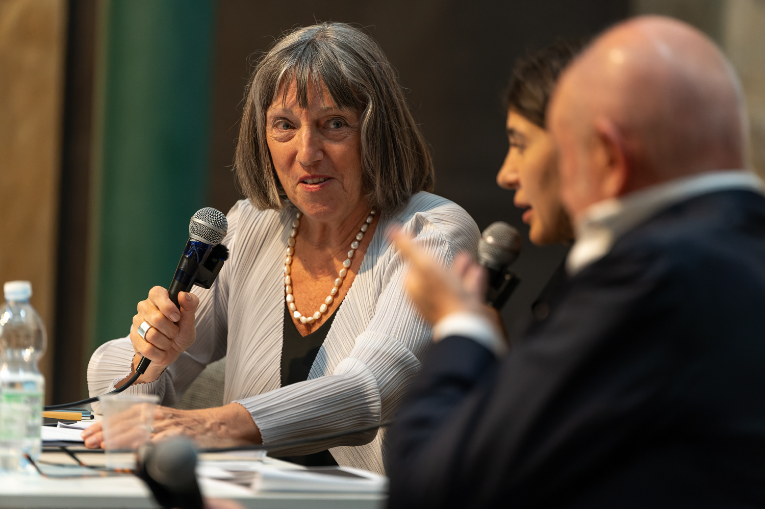 Tre persone discutono con microfoni in mano durante una conferenza. Bottiglia d'acqua sul tavolo.