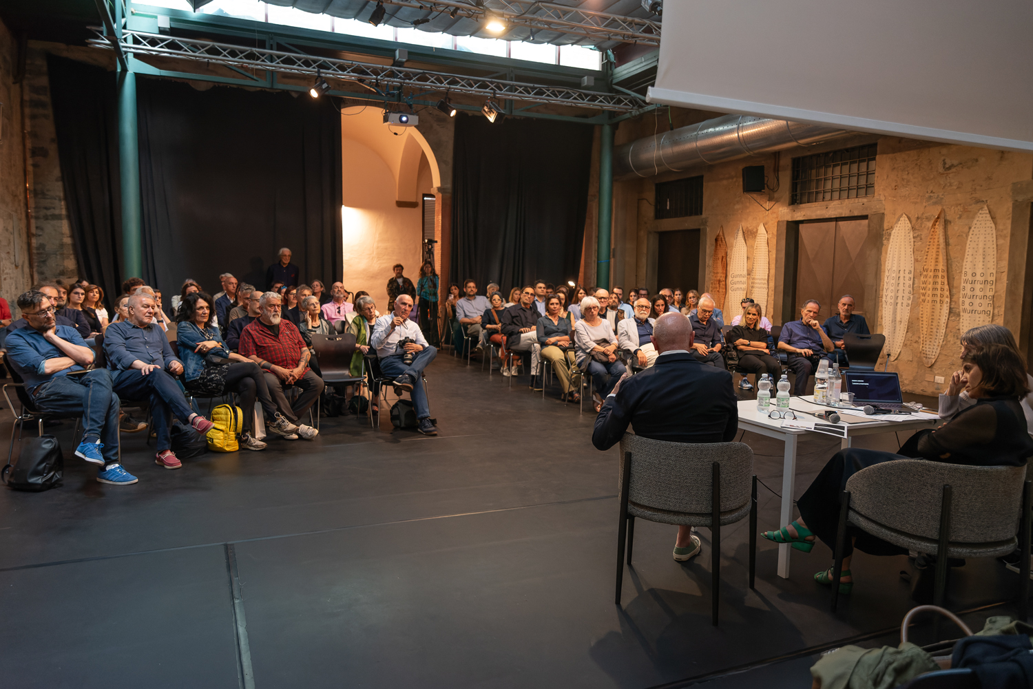 Gruppo di persone partecipa a una conferenza in una sala con drappeggi e attrezzature audiovisive.