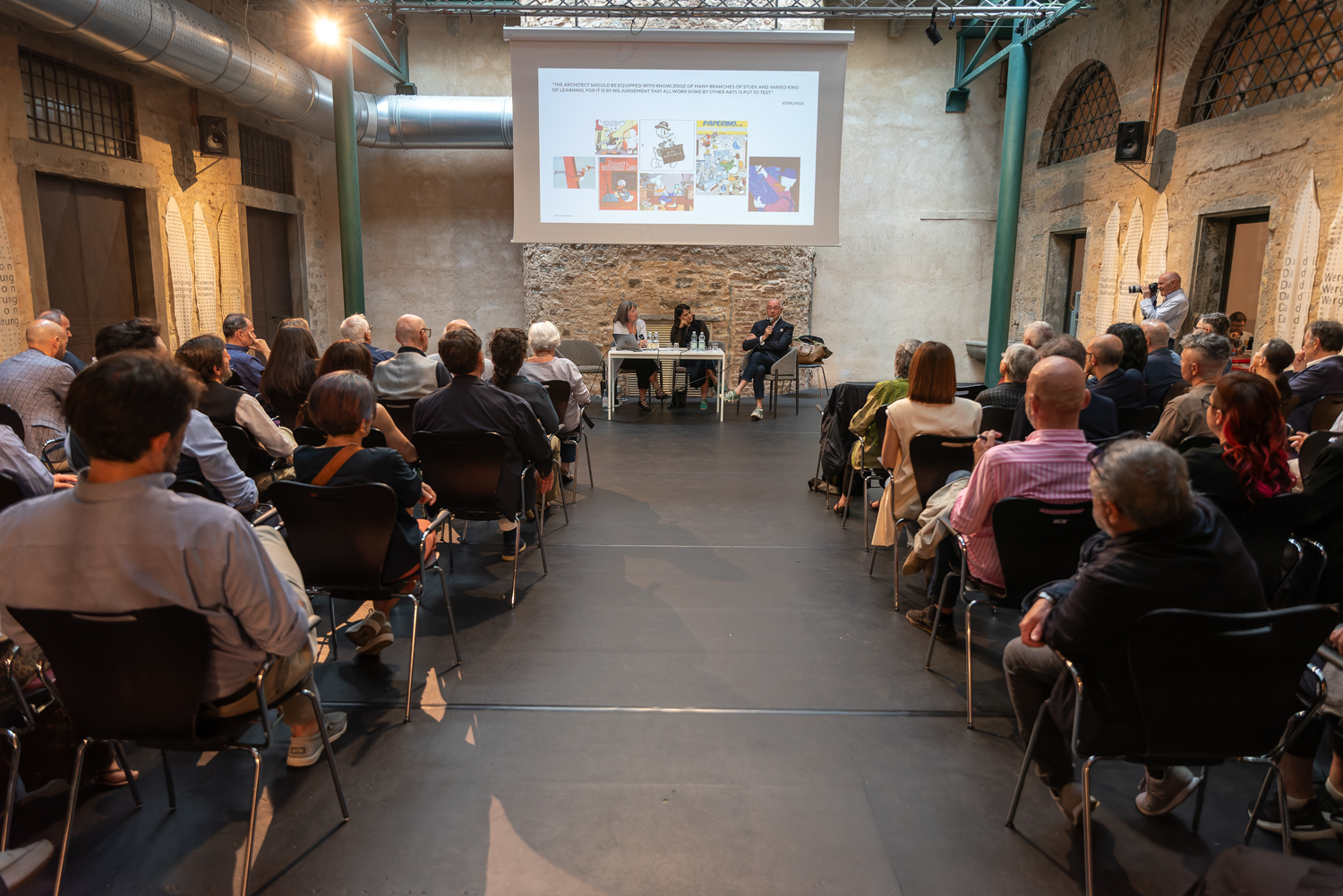 Conferenza in sala conferenze con pubblico attento e tre relatori davanti a un grande schermo proiettore.