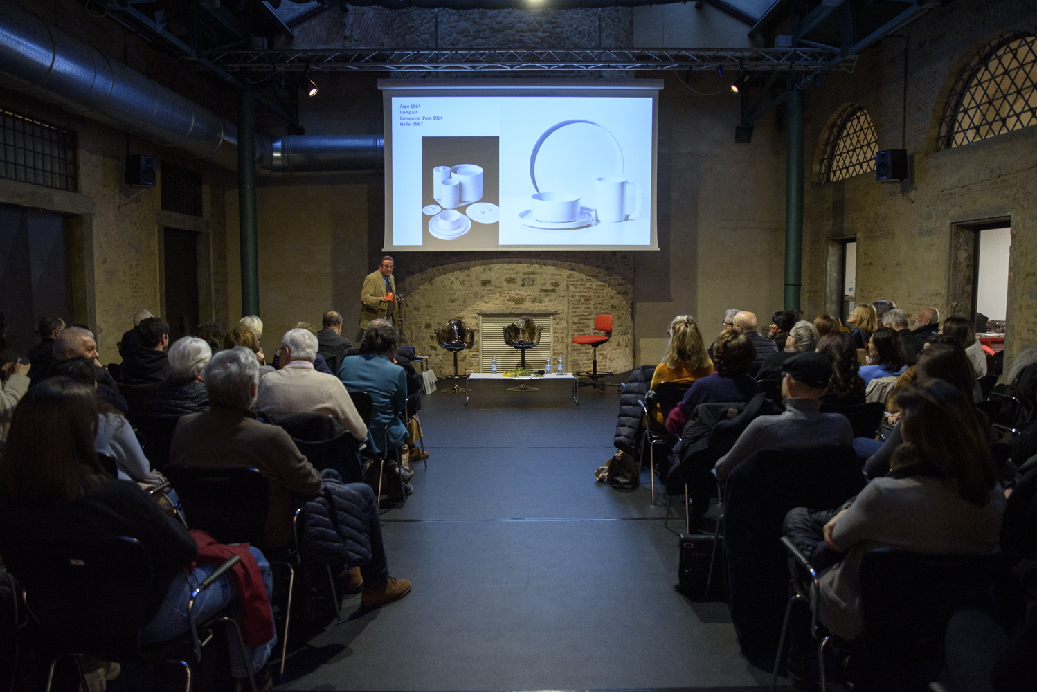 Conferenza su design industriale in sala conferenze con pubblico attento e proiezione di slide su schermo.