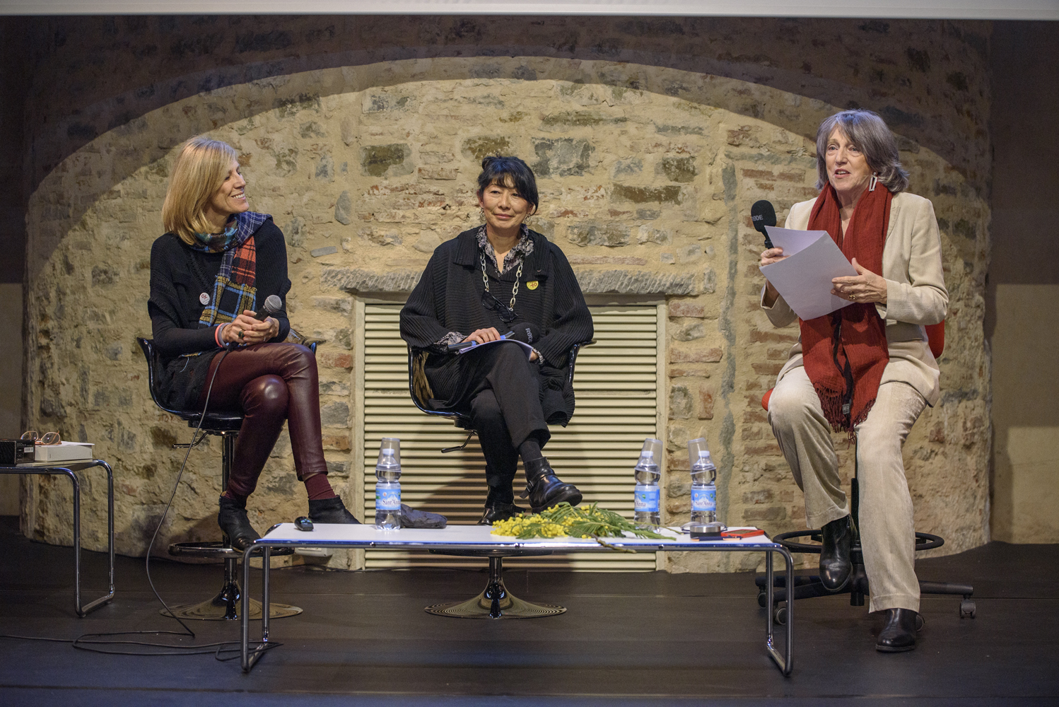 Tre donne siedono su sedie sotto un arco di pietra durante un evento, tenendo microfoni e carte.