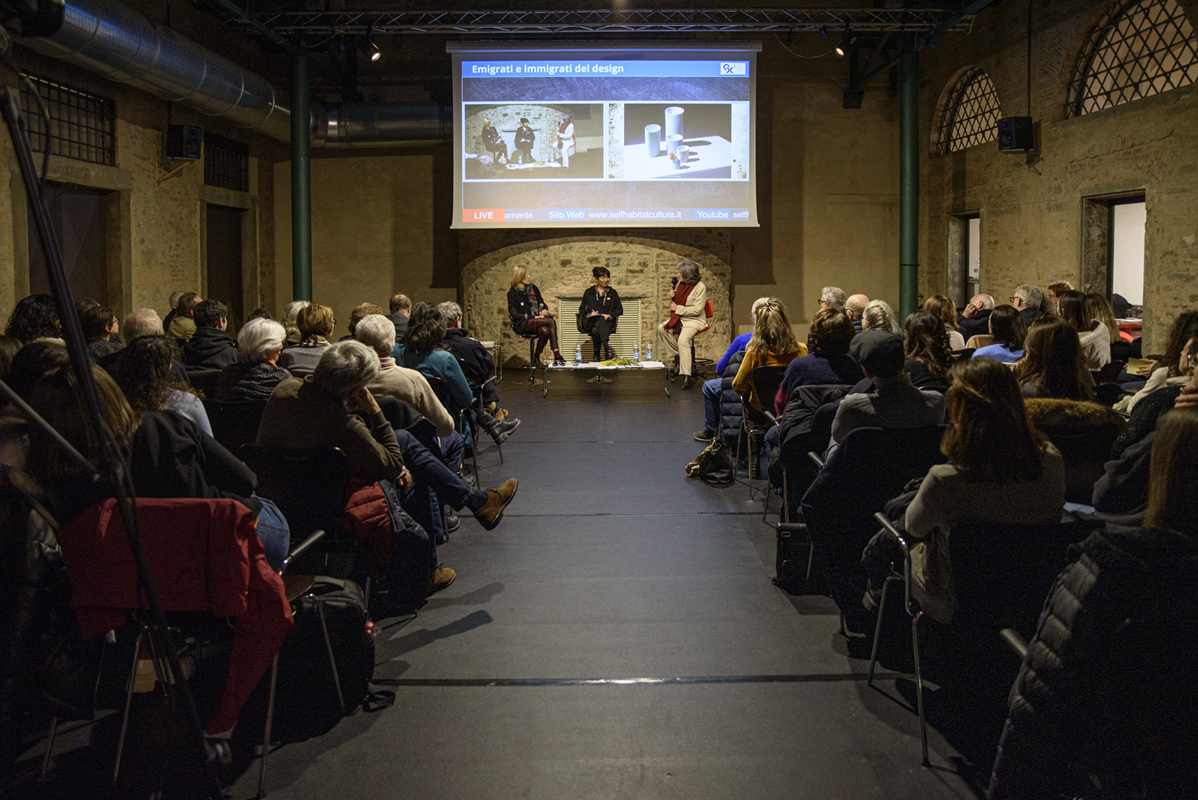 Conferenza su emigrati e design, pubblico attento e relatori sul palco in un ambiente storico.