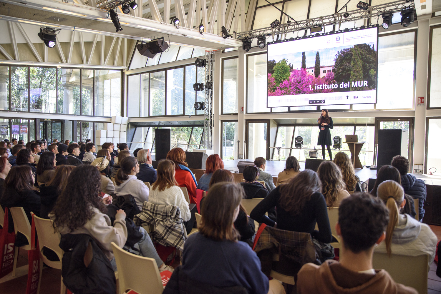 Conferenza in aula moderna con molte persone sedute e una presentazione su grande schermo.