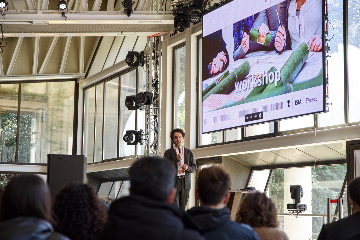 Presentazione in un auditorium con schermo che mostra un workshop, pubblico attento, ambiente moderno e luminoso.