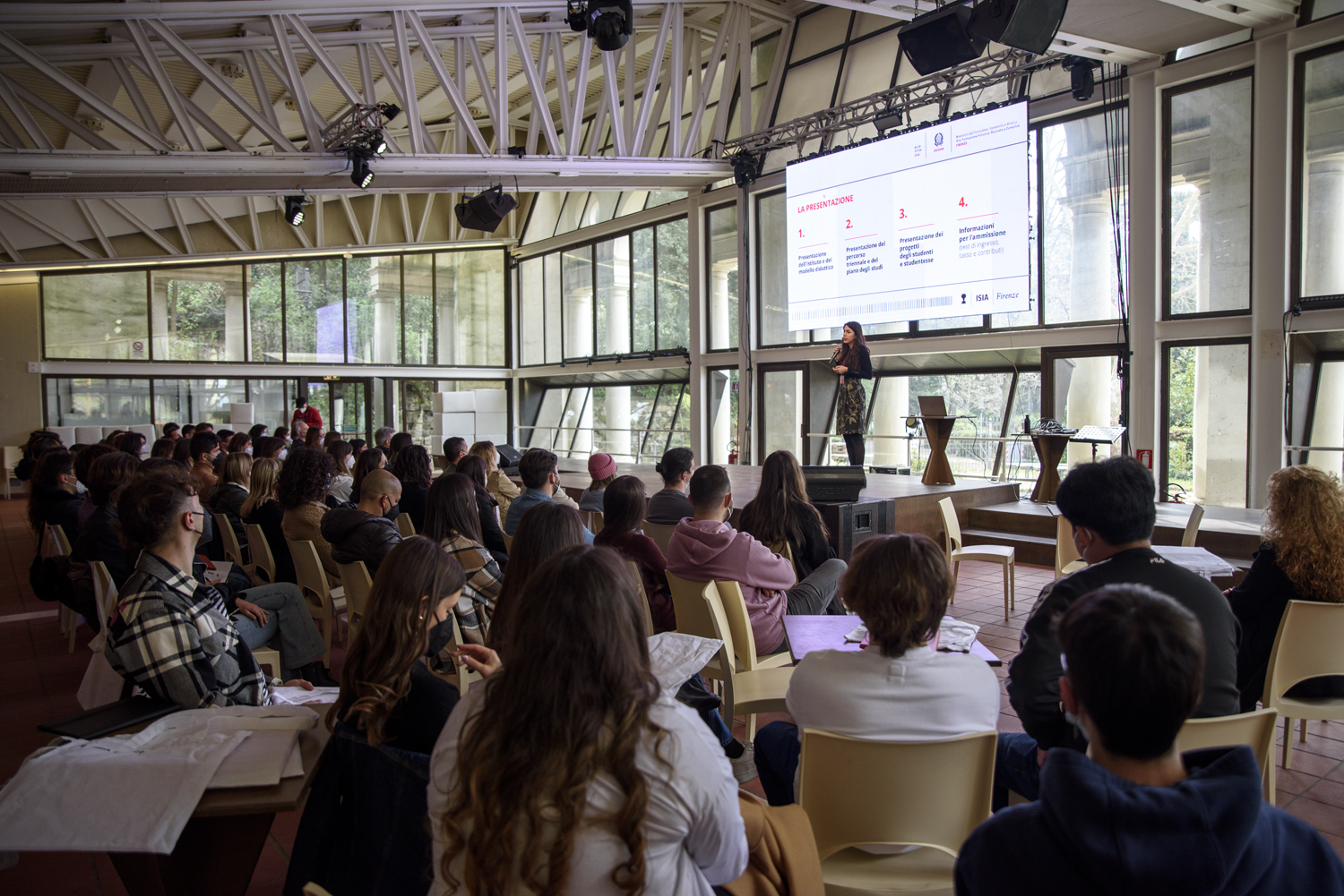 Conferenza in una sala moderna con grandi finestre, relatore sul palco e pubblico attento seduto.