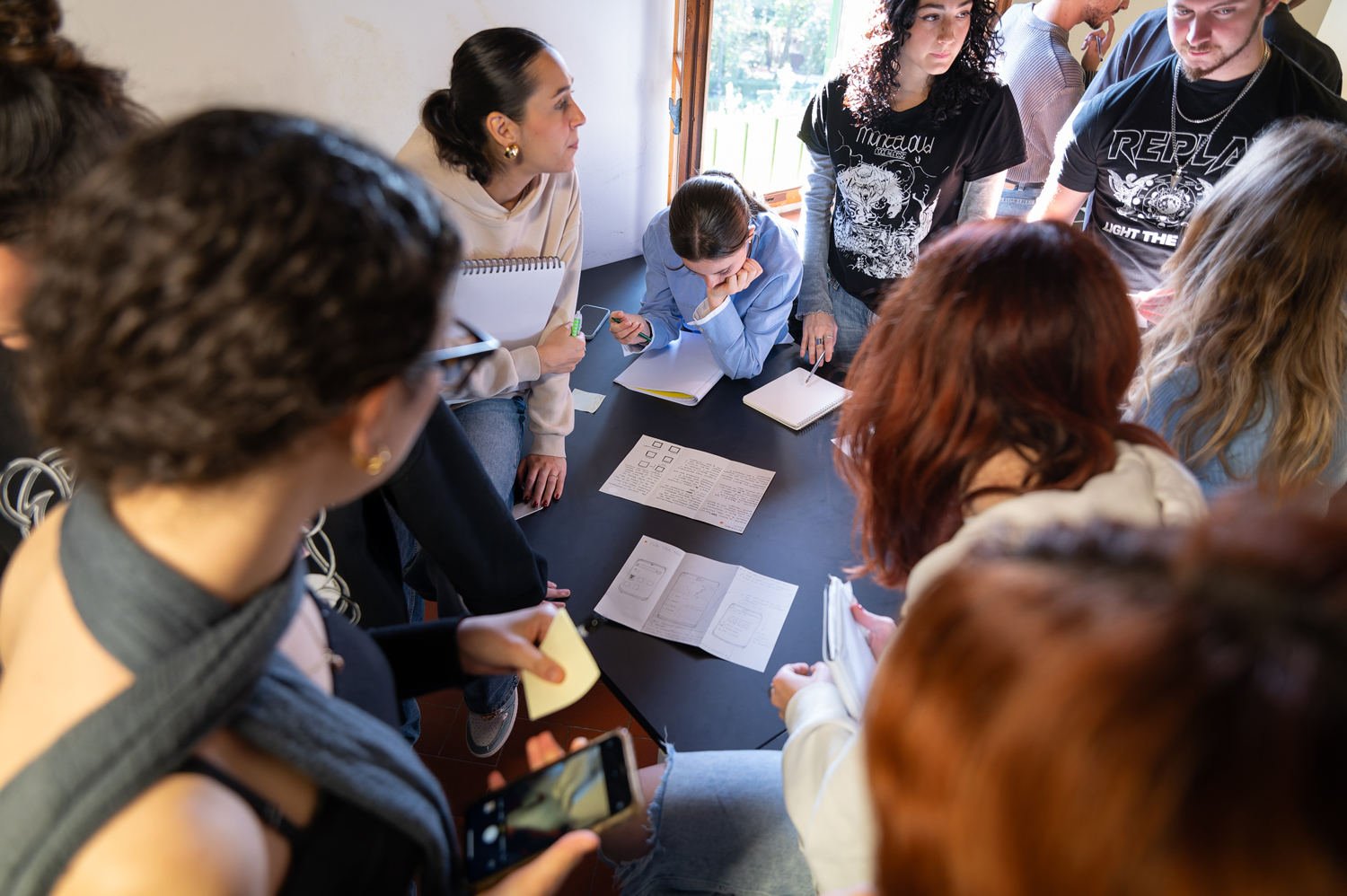 Gruppo di persone in una riunione, discutendo e prendendo appunti intorno a un tavolo, con quaderni e smartphone.