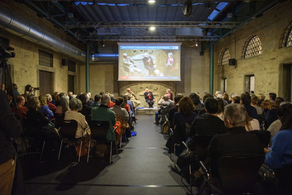 Un pubblico ascolta una conferenza con un relatore, in un ambiente rustico con schermo proiettato sullo sfondo.