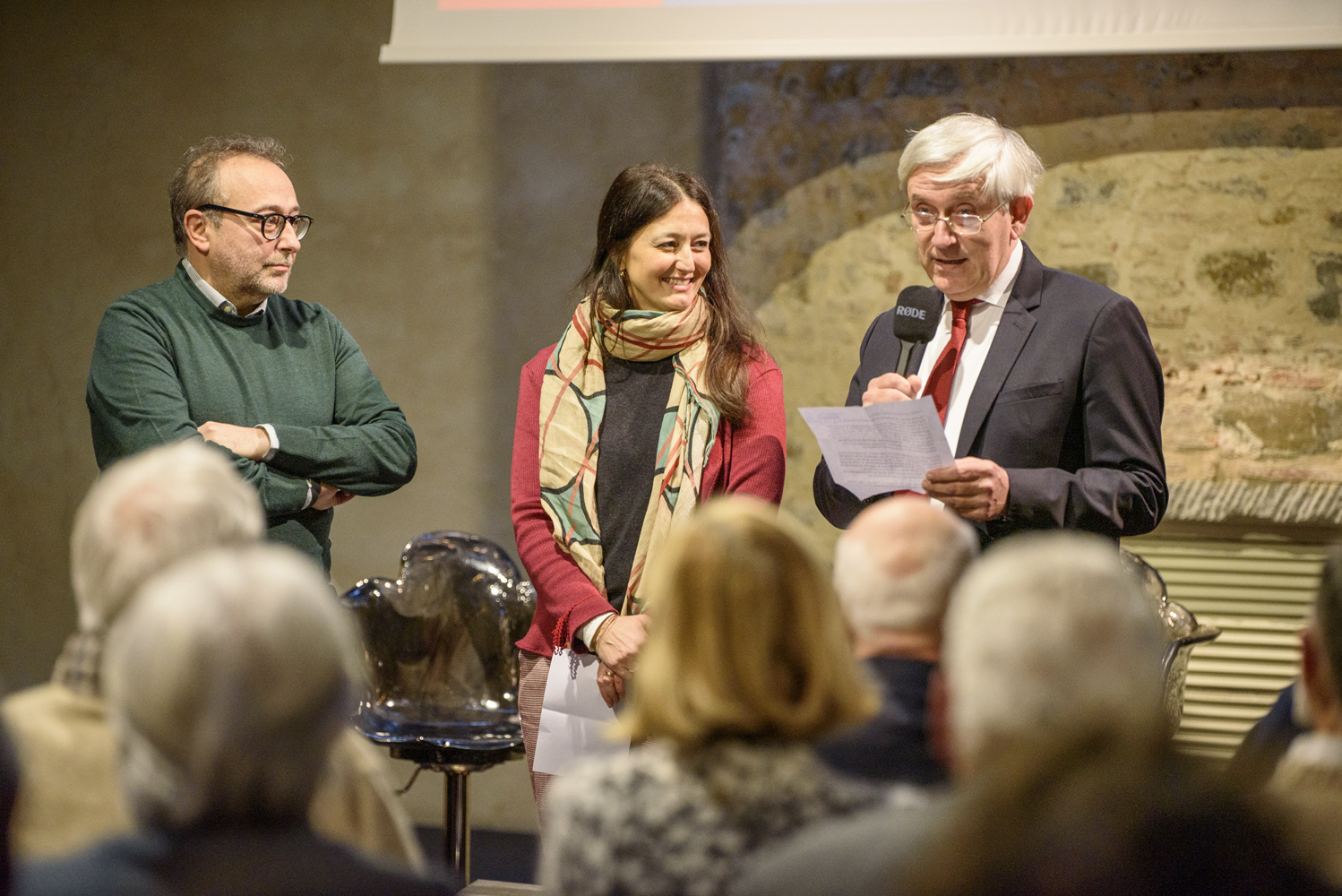 Tre persone parlano durante un evento con pubblico attento in una sala con pareti in pietra.
