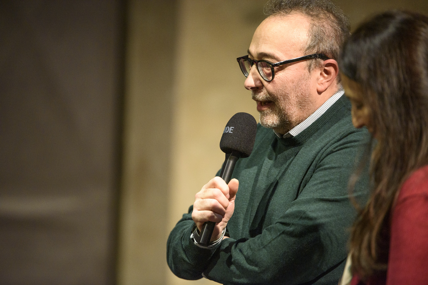 Uomo con occhiali parla al microfono durante un evento, con una donna accanto a lui sul palco.