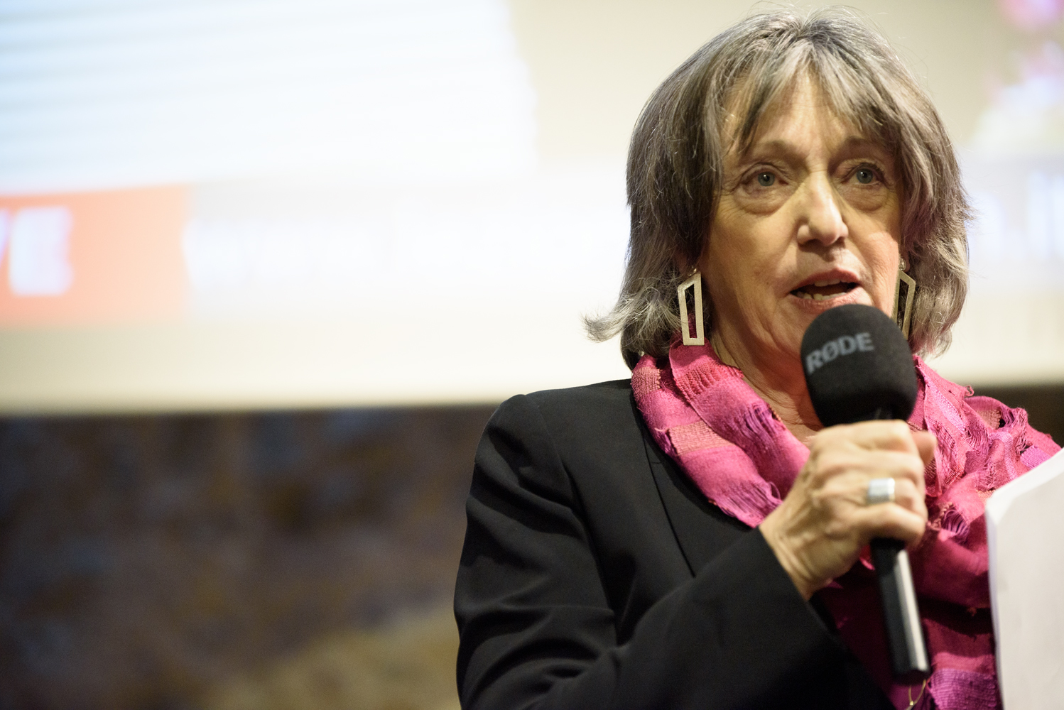 Persona anziana parla al microfono durante un evento, indossando una sciarpa rosa e orecchini geometrici.
