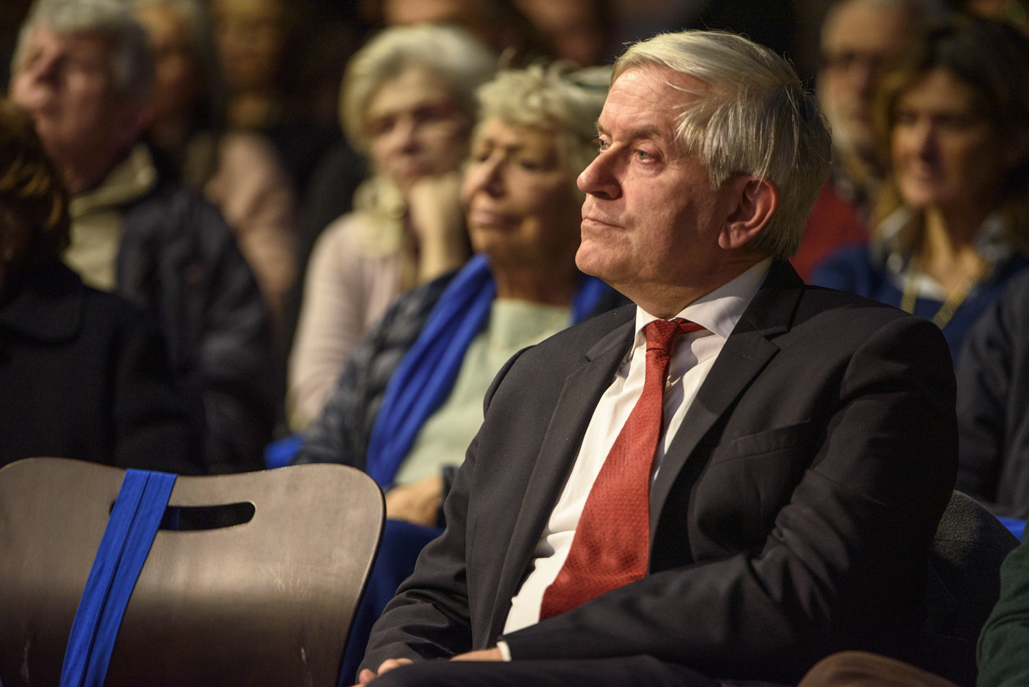 Un uomo in abito elegante ascolta con attenzione in una platea piena di persone durante un evento pubblico.