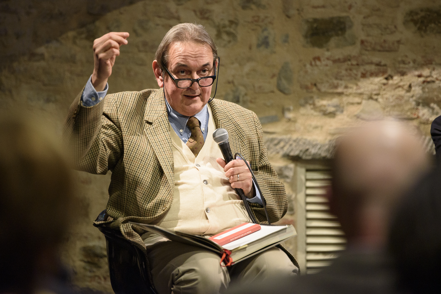 Persona anziana discute in pubblico con microfono e libro, indossa occhiali e abito elegante, sfondo di mattoni.