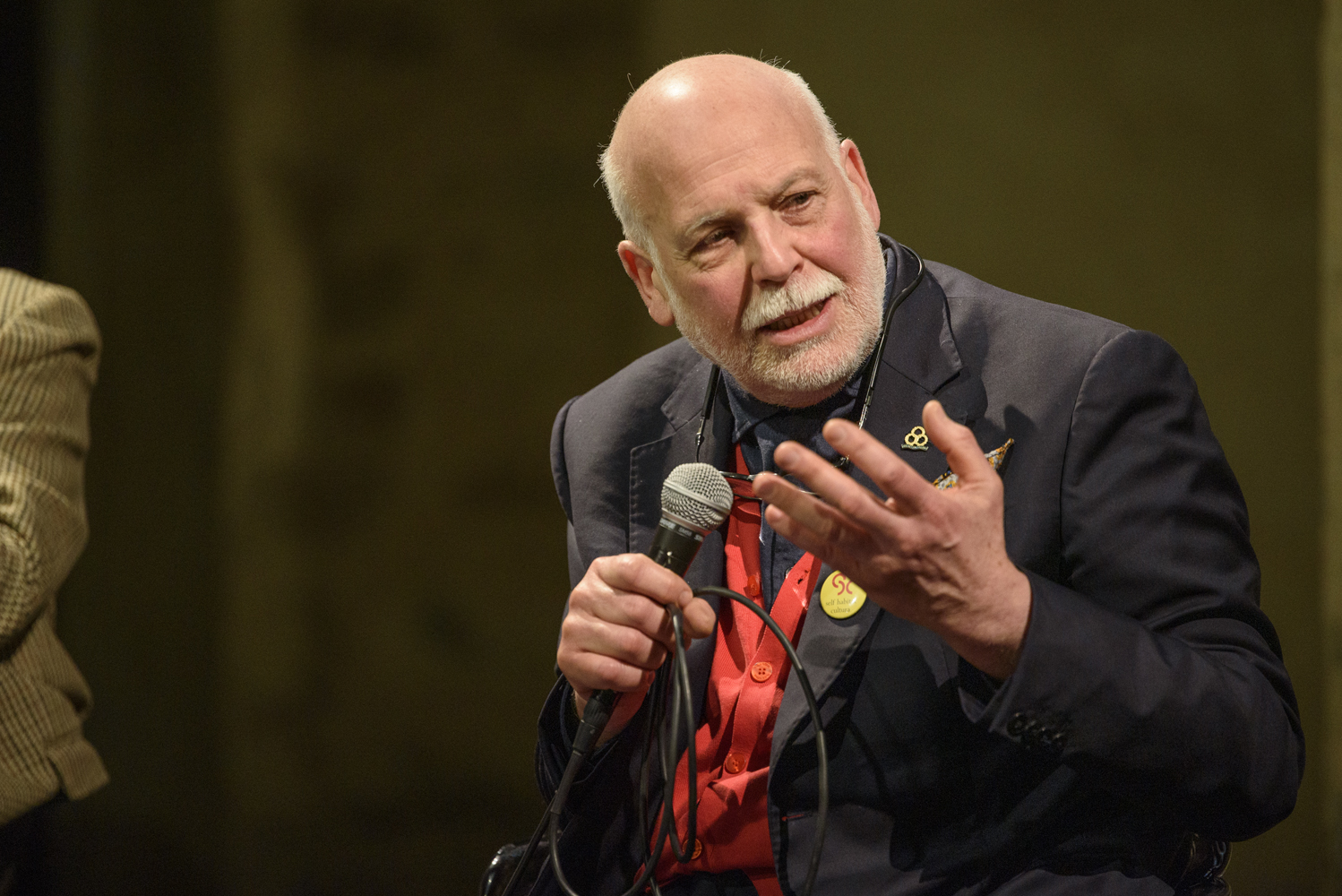 Uomo anziano con microfono, abito scuro e camicia rossa, discute animatamente in un evento pubblico.