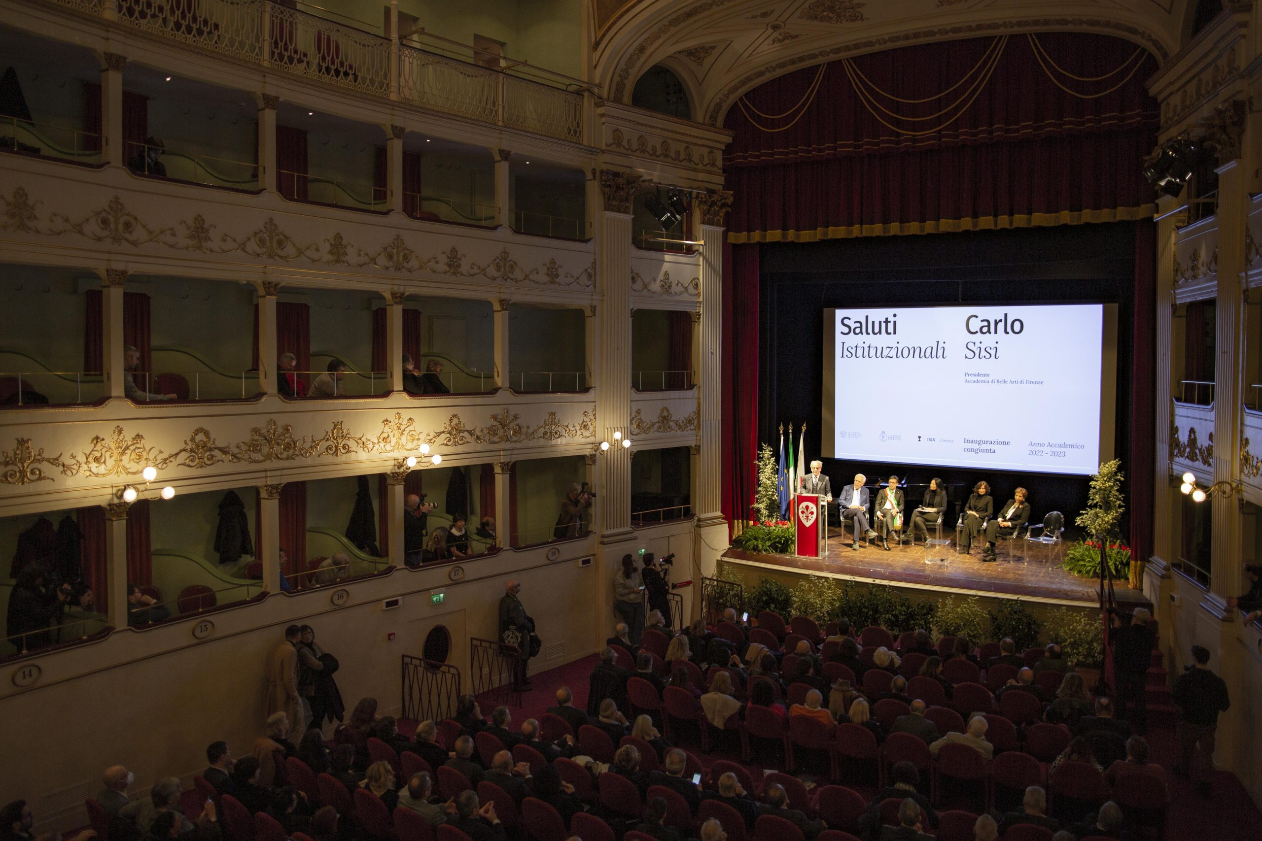 Inaugurazione accademica in teatro storico con pubblico e ospiti su palco, proiezione saluti istituzionali.