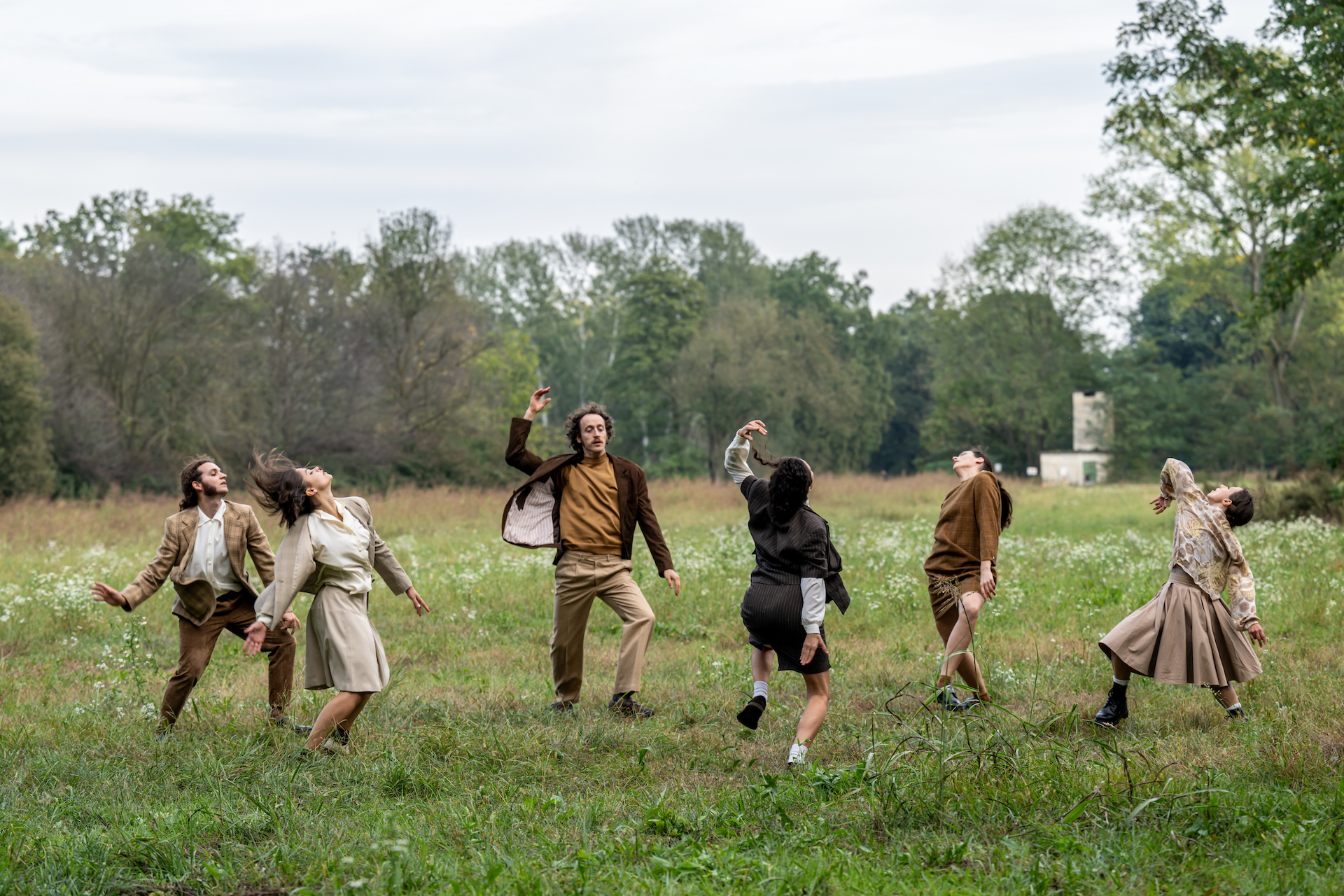 Ballerini in abiti casual danzano in un campo verde, circondati da alberi e cielo nuvoloso.